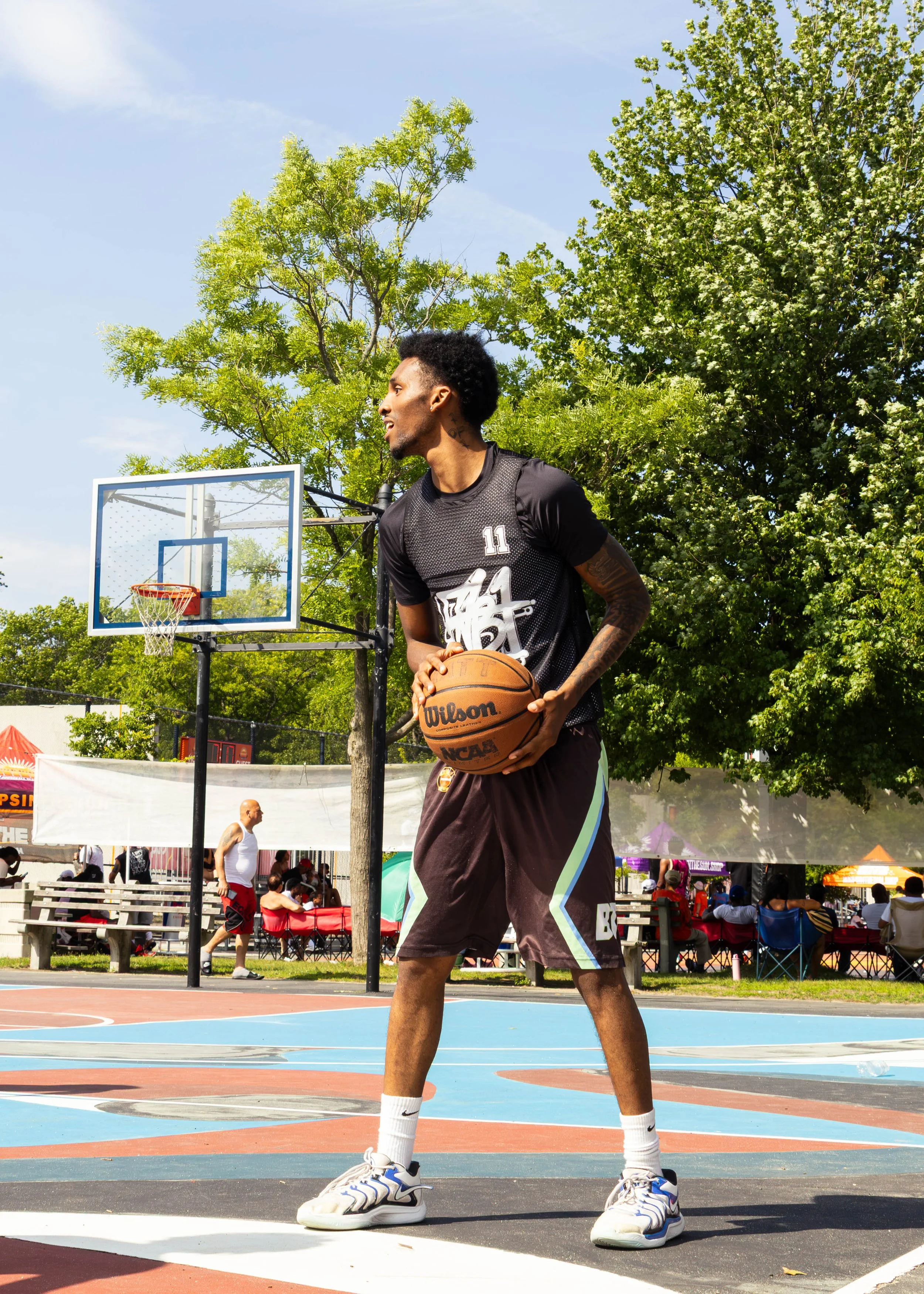 Young man with curly hair wearing a basketball jersey, shorts, and sneakers stands on an outdoor basketball court holding a basketball. There are green trees, a crowd of people, and a basketball hoop in the background on a sunny day.