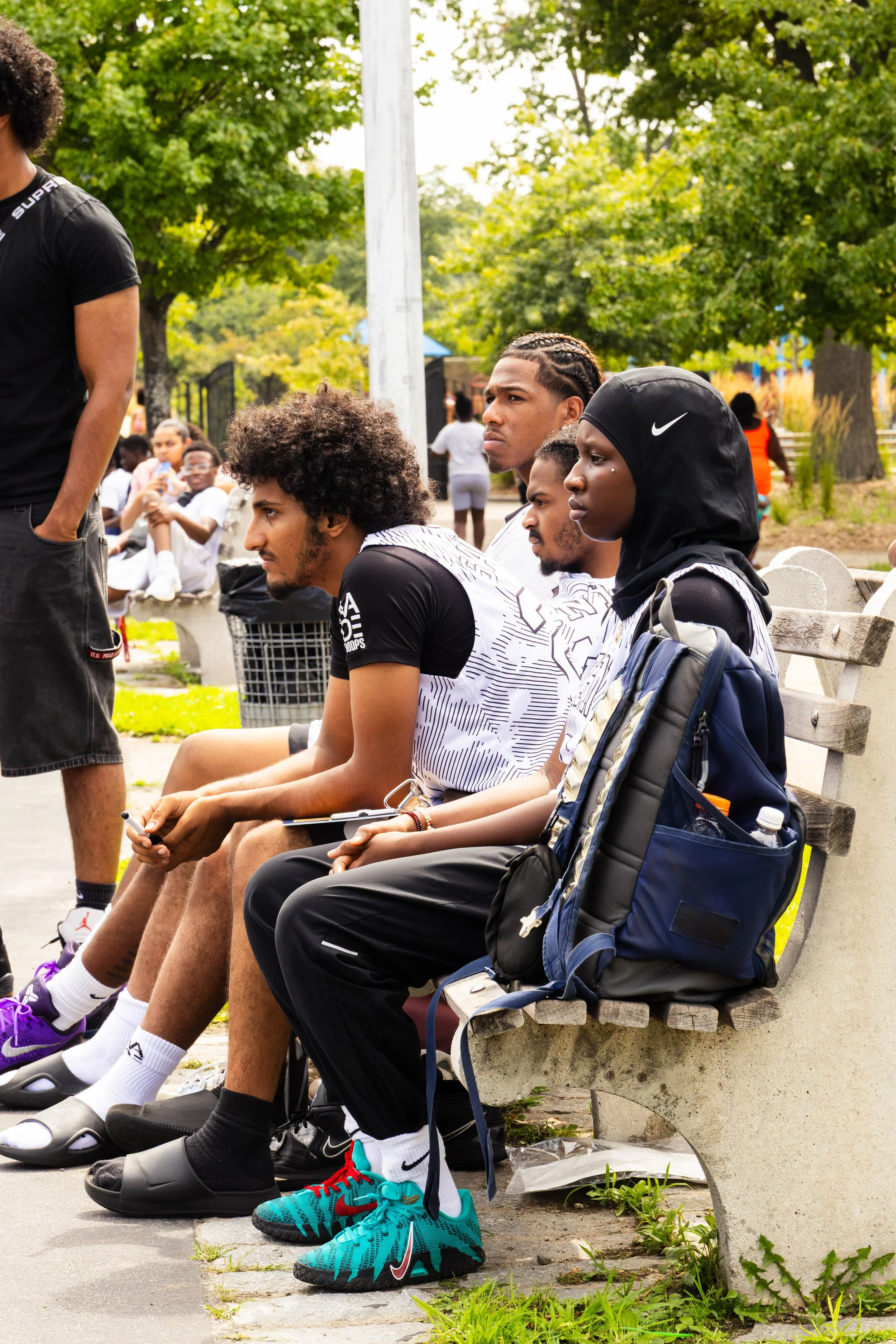 People sitting on a park bench, listening attentively, with trees and greenery in the background. They are wearing athletic clothing and sneakers.