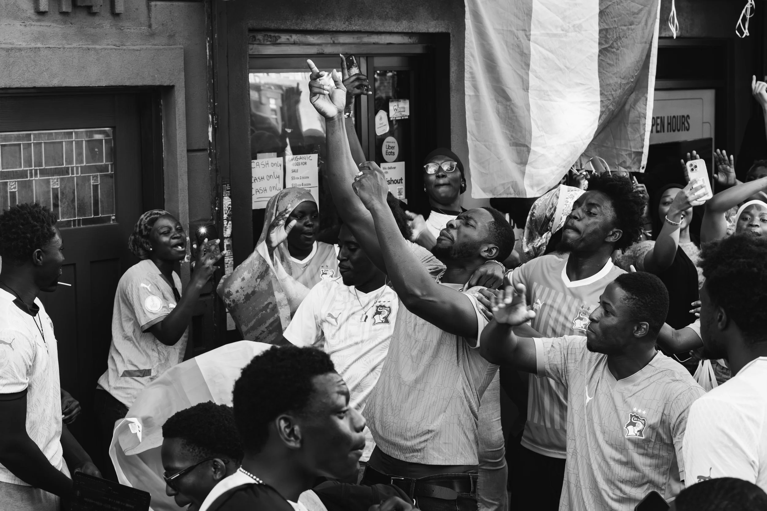 A group of people gathered outdoors, with some raising their hands and others taking photos or videos. The scene appears lively and celebratory.