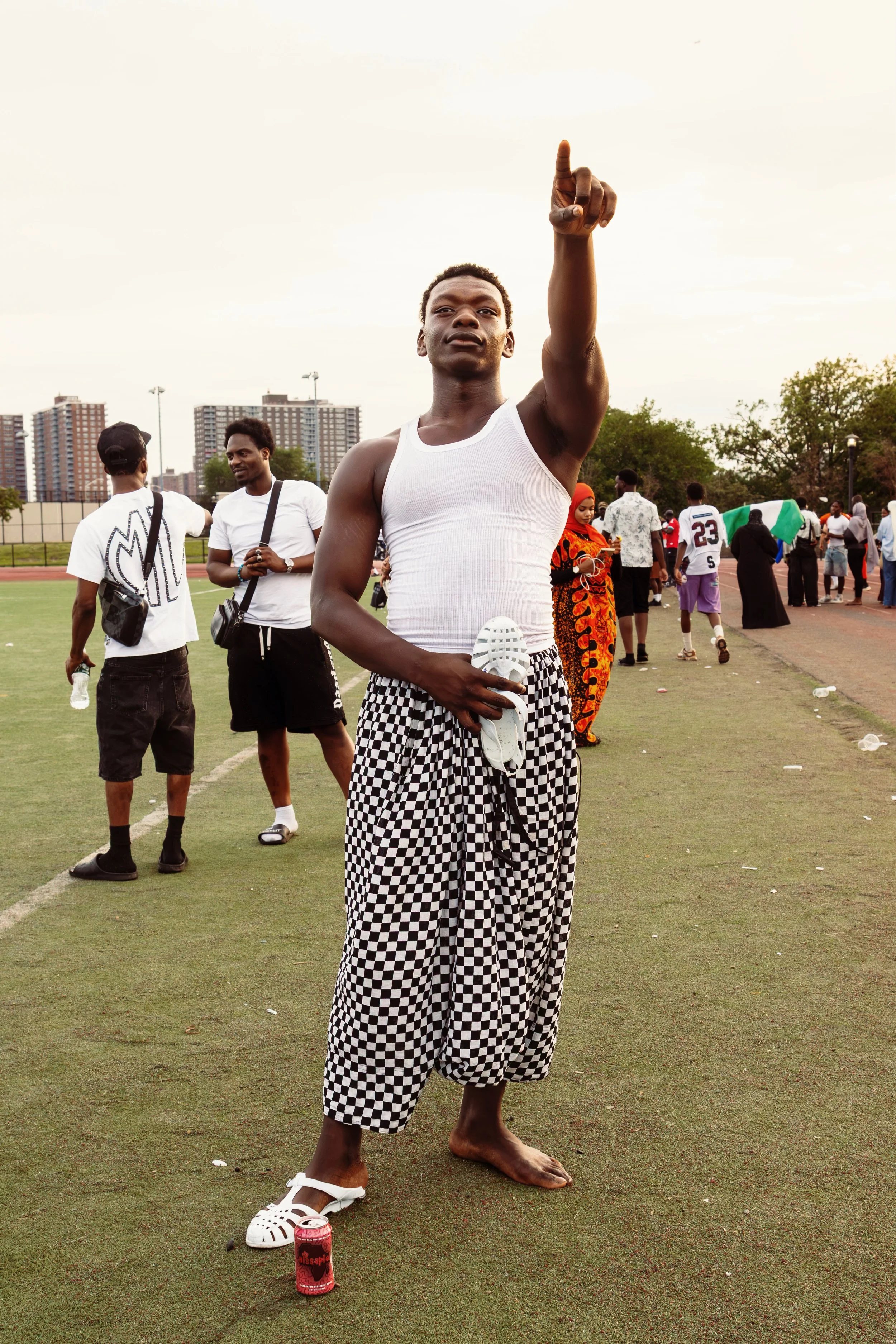 A young man with dark skin and short curly hair dressed in a white tank top and checkered pants stands barefoot on a sports field. He is pointing upward with his right hand, holding a white shoe in his left hand, and standing next to a red soda can. 