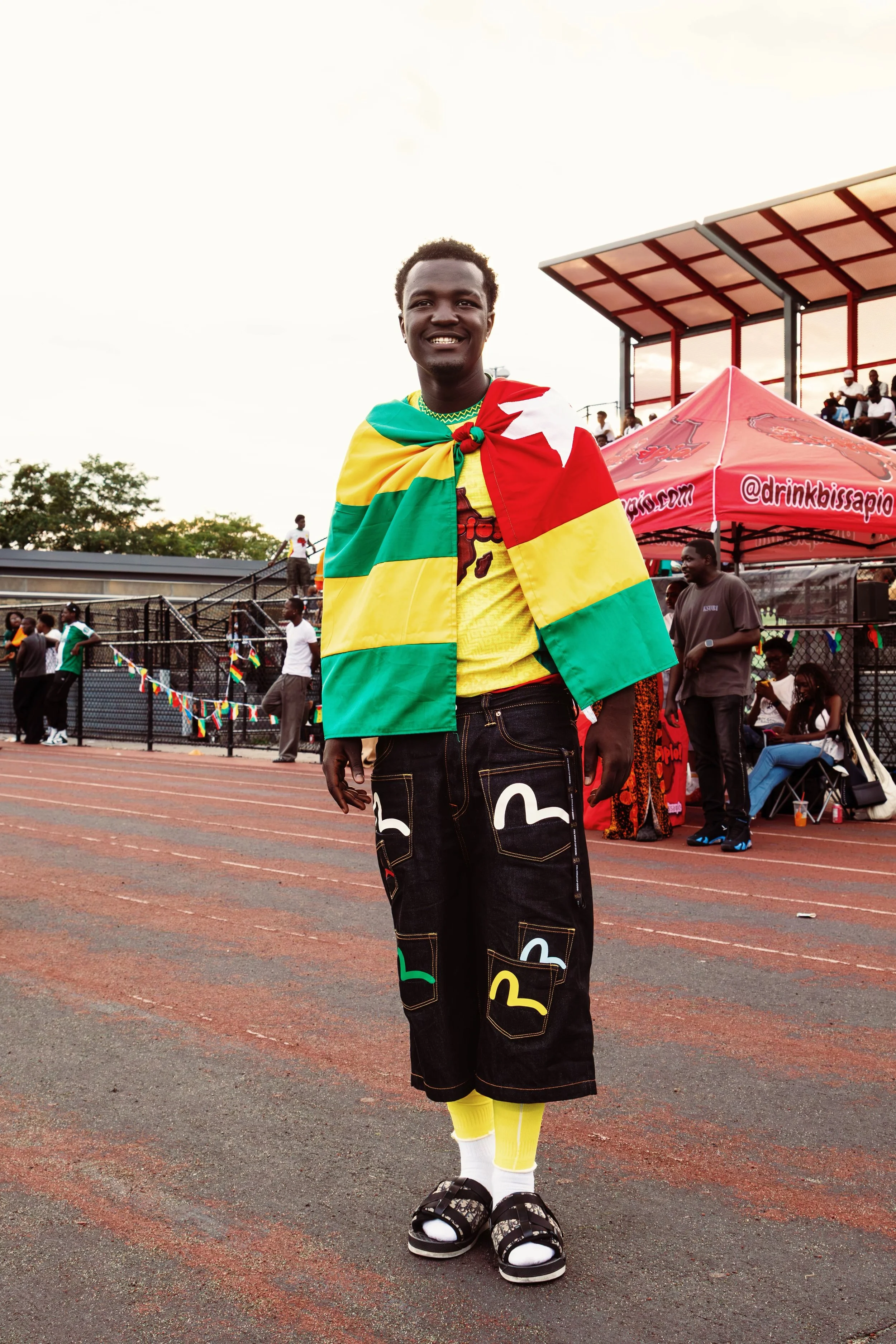 A young man smiling, wearing a yellow shirt, black shorts with colorful patches, white and yellow socks, black sandals, and draped in a multicolored flag of green, yellow, red, and white. He is standing on an outdoor track with spectators and a red c