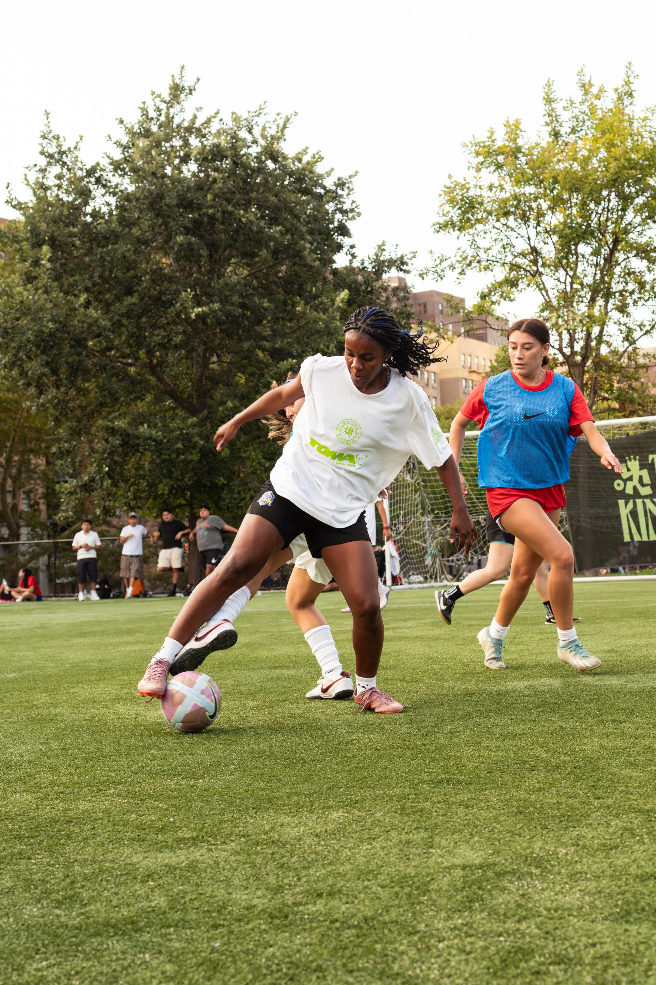 Two female soccer players competing for the ball on a grassy field during a match, with spectators and trees in the background.