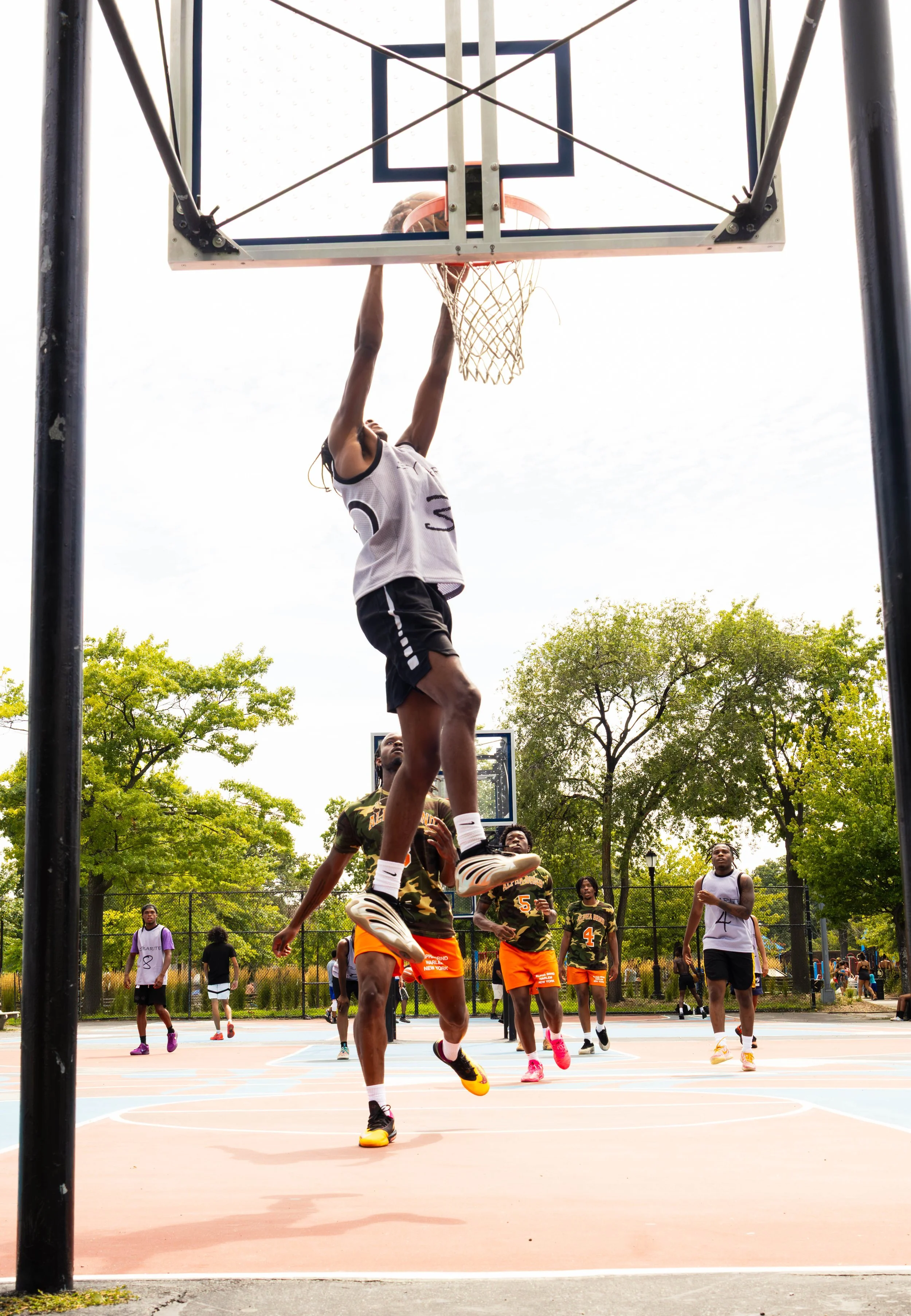 A basketball game on an outdoor court, with one player attempting to make a dunk and others watching or preparing to play, surrounded by trees and a fence.