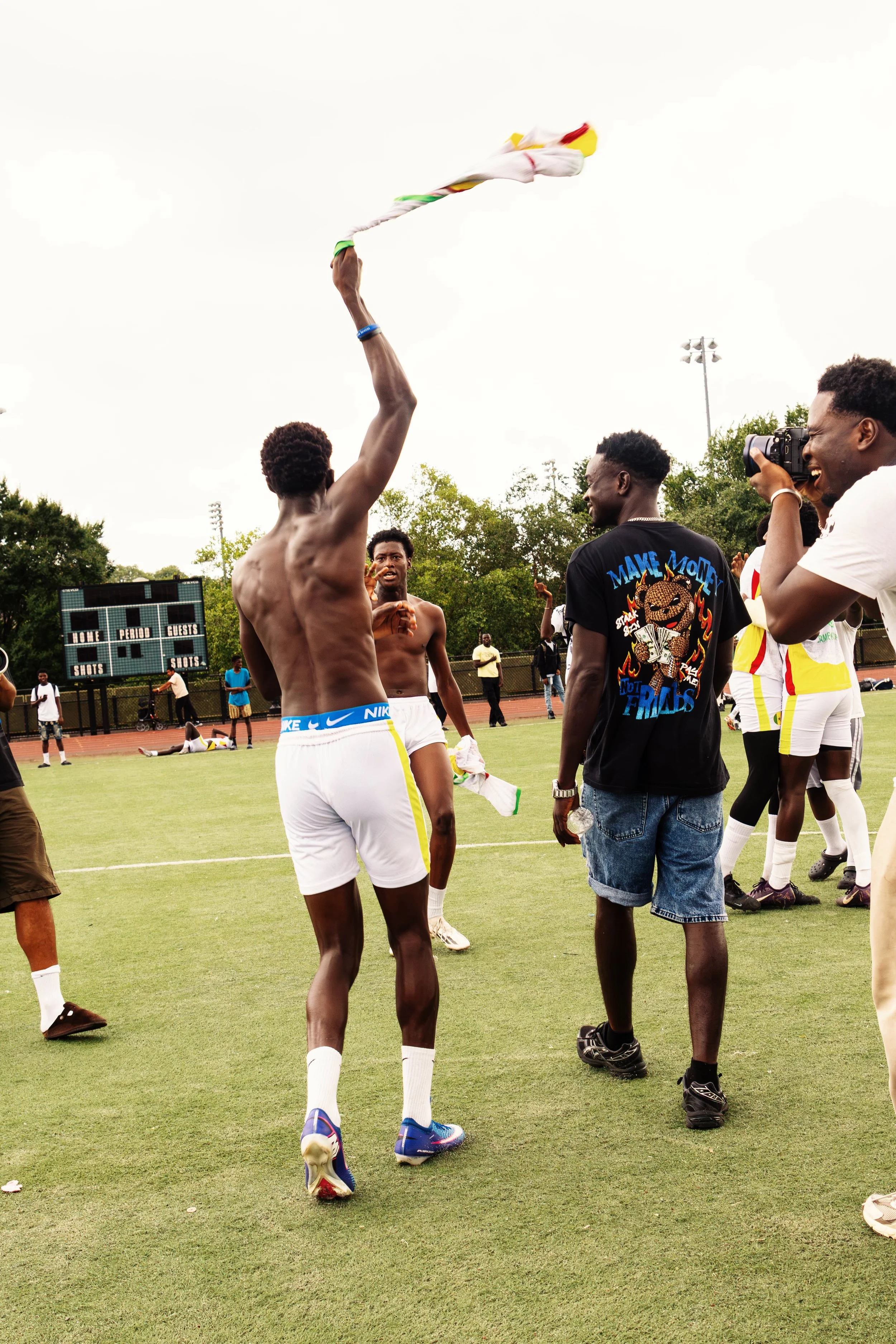 Group of young men on a sports field, some are shirtless, one holding a flag, others are taking photos or talking, with a scoreboard in the background.