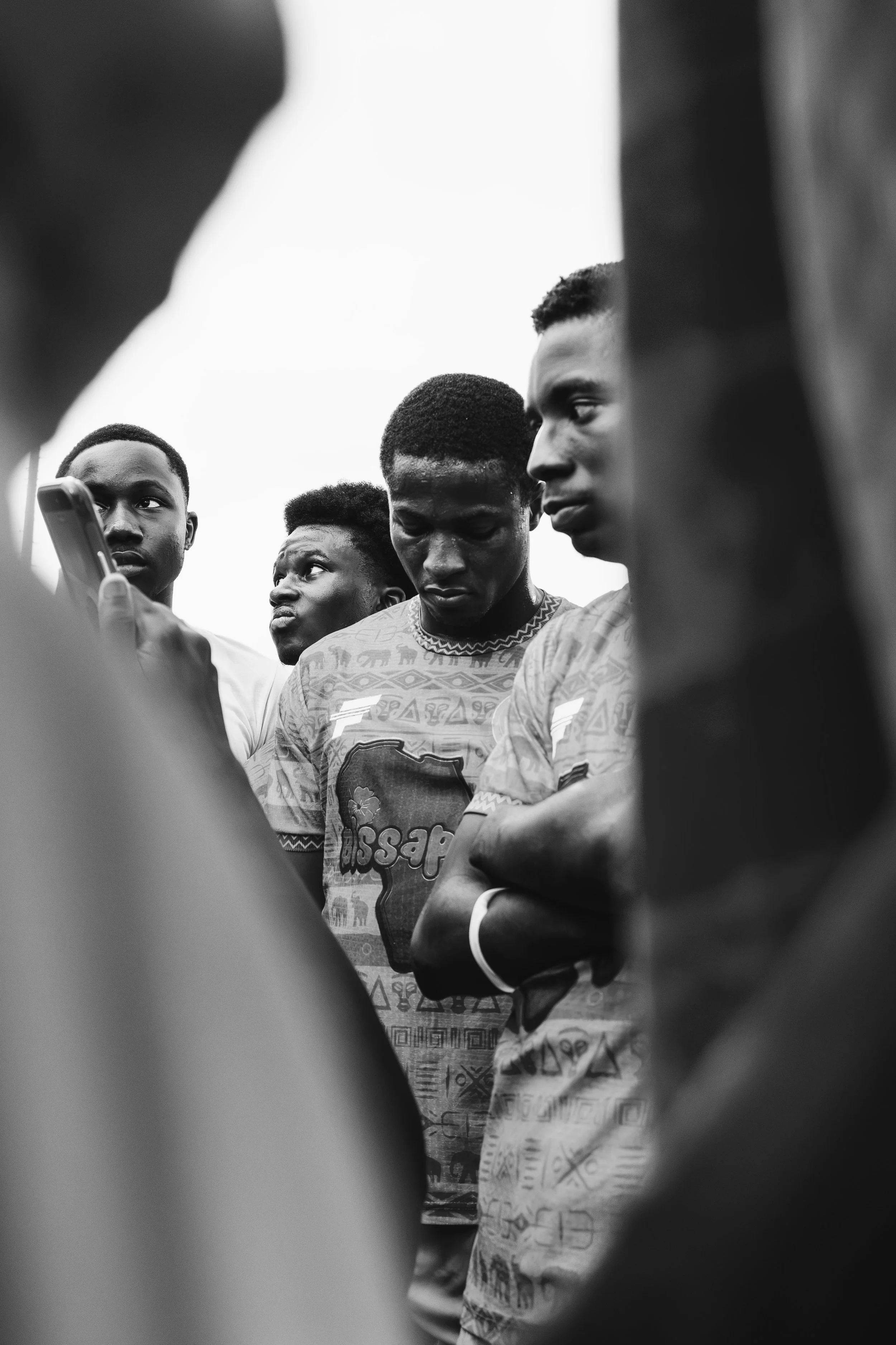 Black and white photograph of four young men standing together, some looking down while others look off into the distance. The image is framed in an unbalanced way with partial views of other people or objects surrounding them.