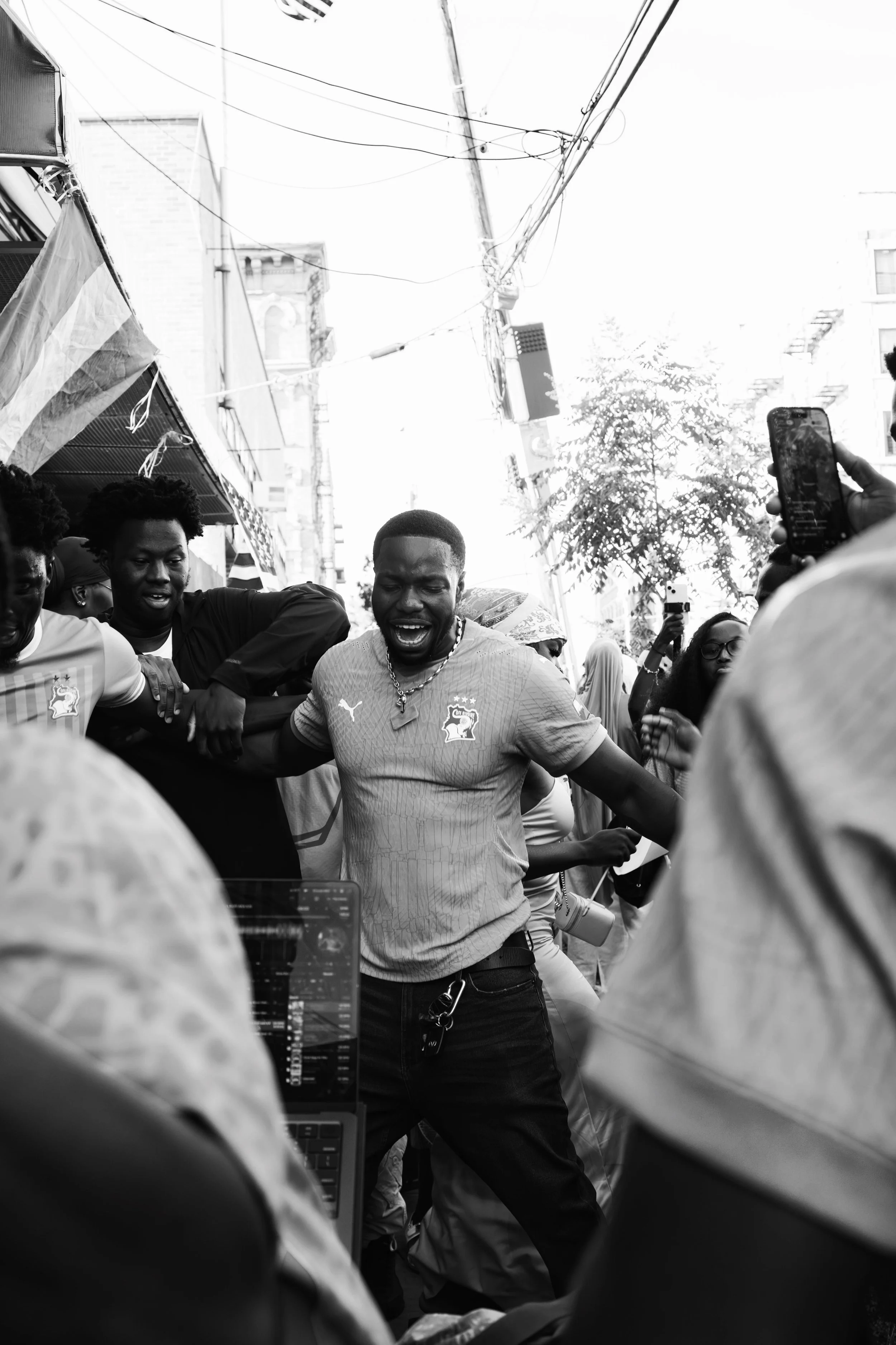 A group of people, including a man in a soccer jersey, gather outdoors on a city street. Some are taking photos or videos, and there are various flags and overhead power lines visible. The scene appears lively and energetic.