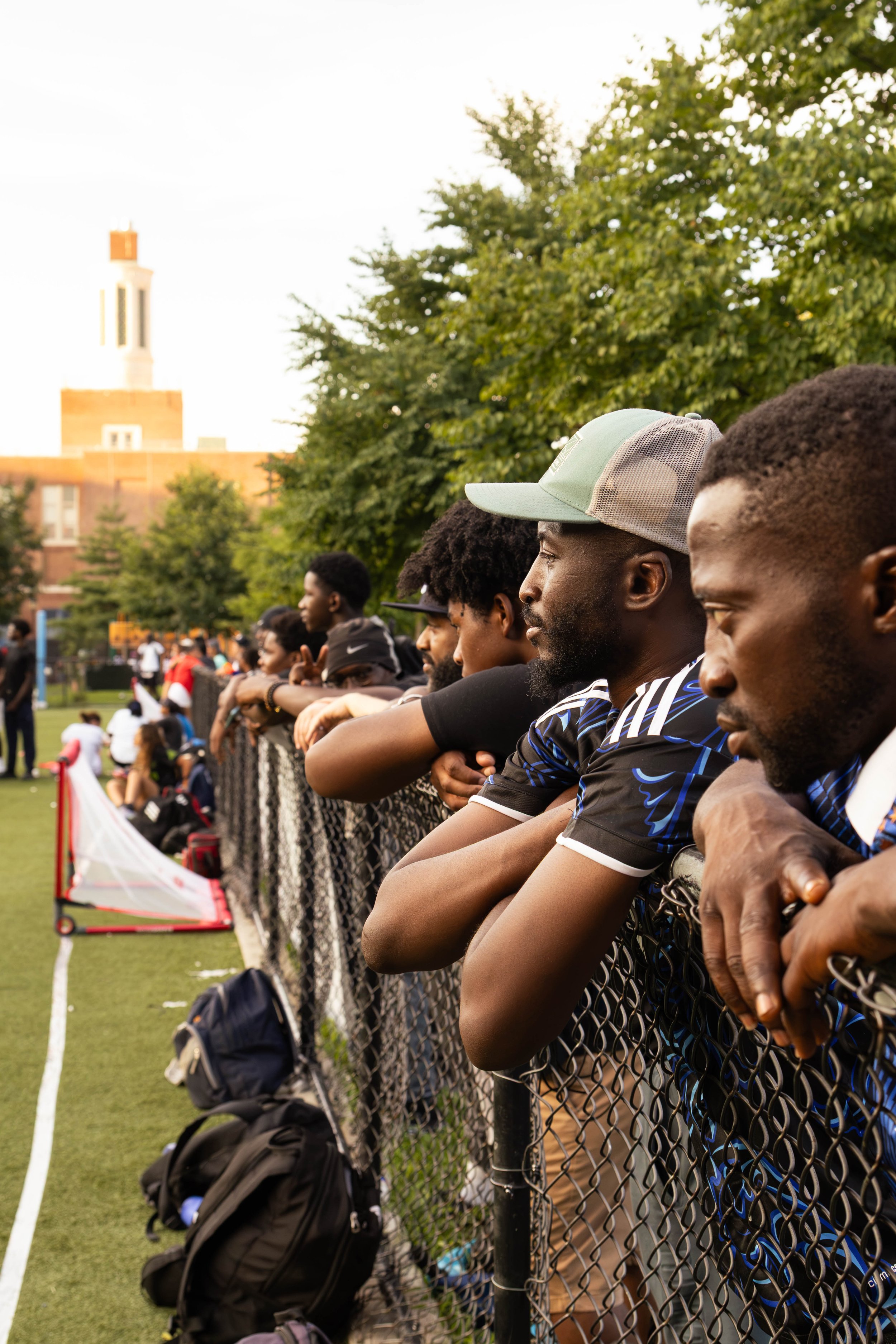 Group of young men standing along a chain-link fence on a sports field, with trees and a building in the background, during daytime.