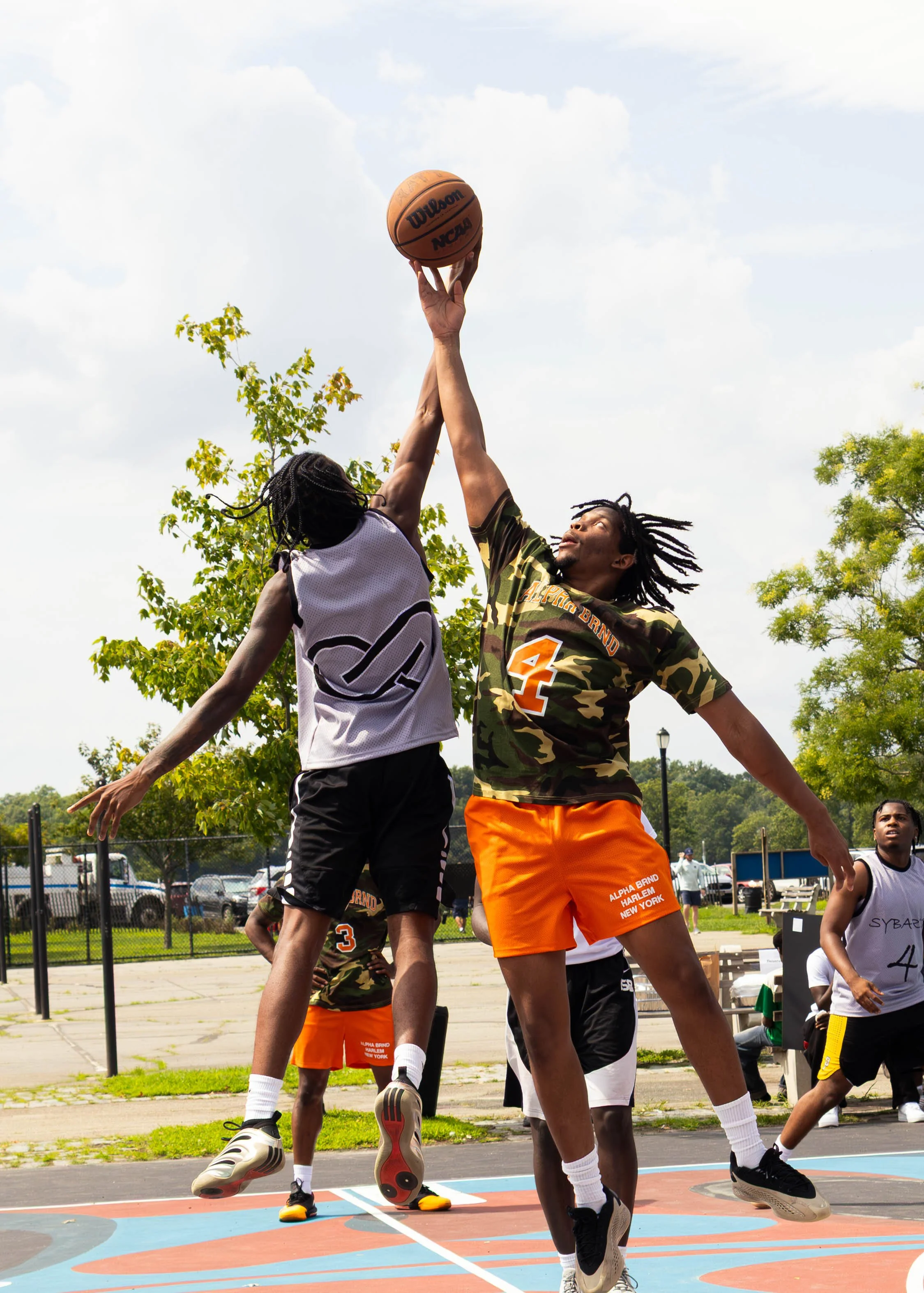 Two basketball players, one in a white jersey and one in a camouflage shirt, jump to contest a shot on an outdoor basketball court. Trees and people are visible in the background.