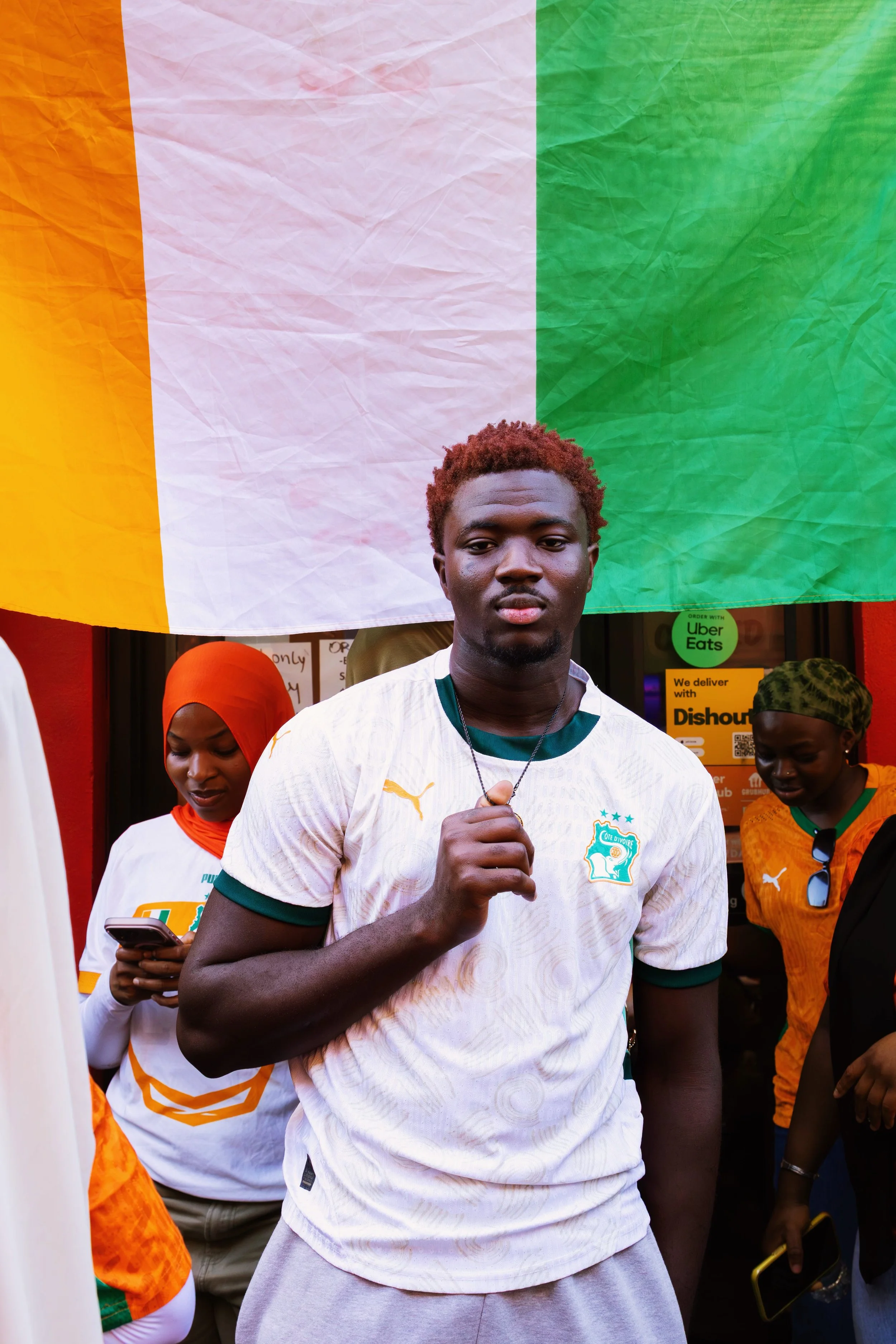 A young man with short red hair wearing a white sports jersey with green accents, standing in front of an Irish flag and a group of people, some of whom are using their phones and wearing sports jerseys.