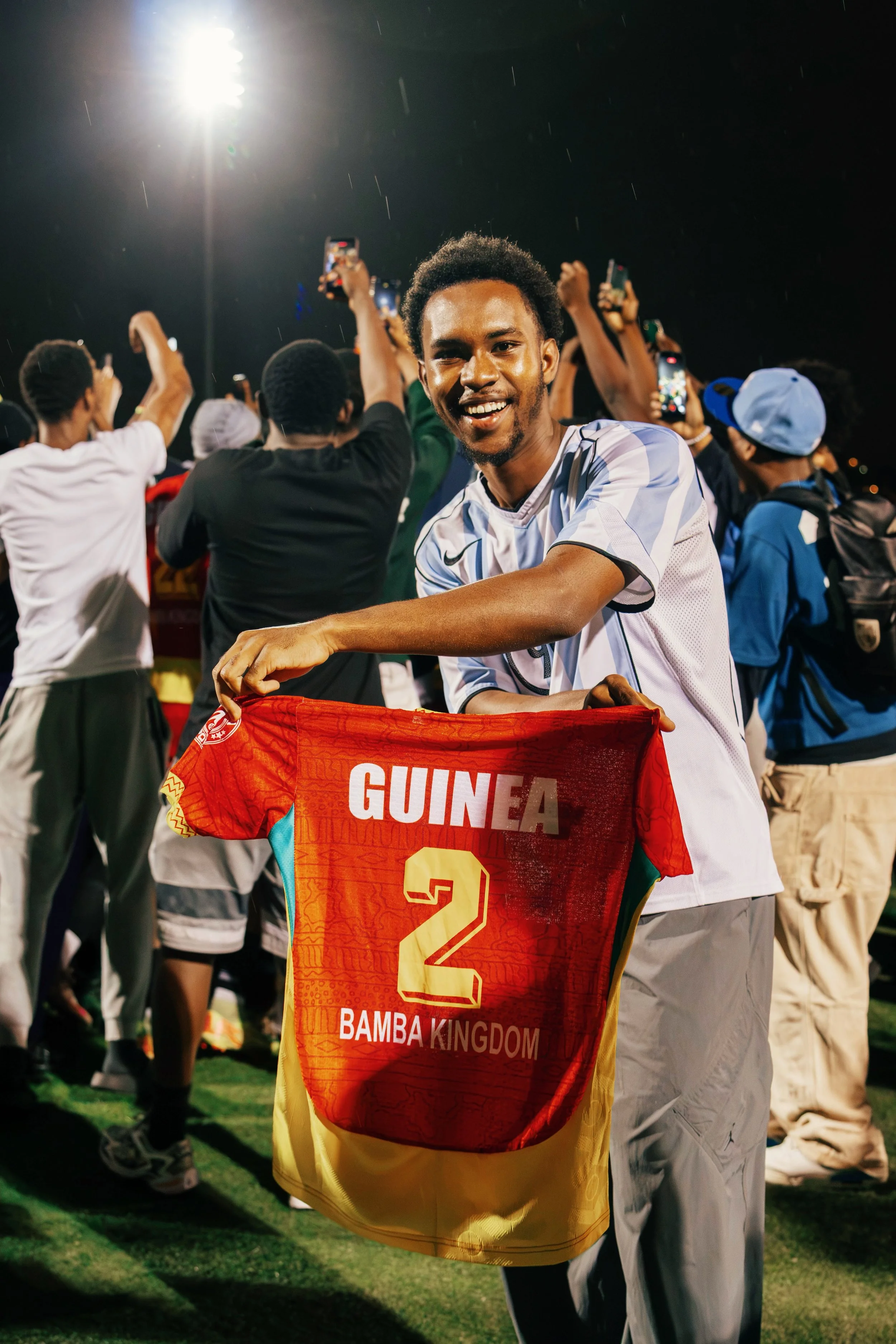 A young man smiling at a celebration, holding a red and yellow scarf with the words 'Guinea Bamb Kingdom' and number 2, surrounded by a crowd taking photos and videos at night under bright lights.
