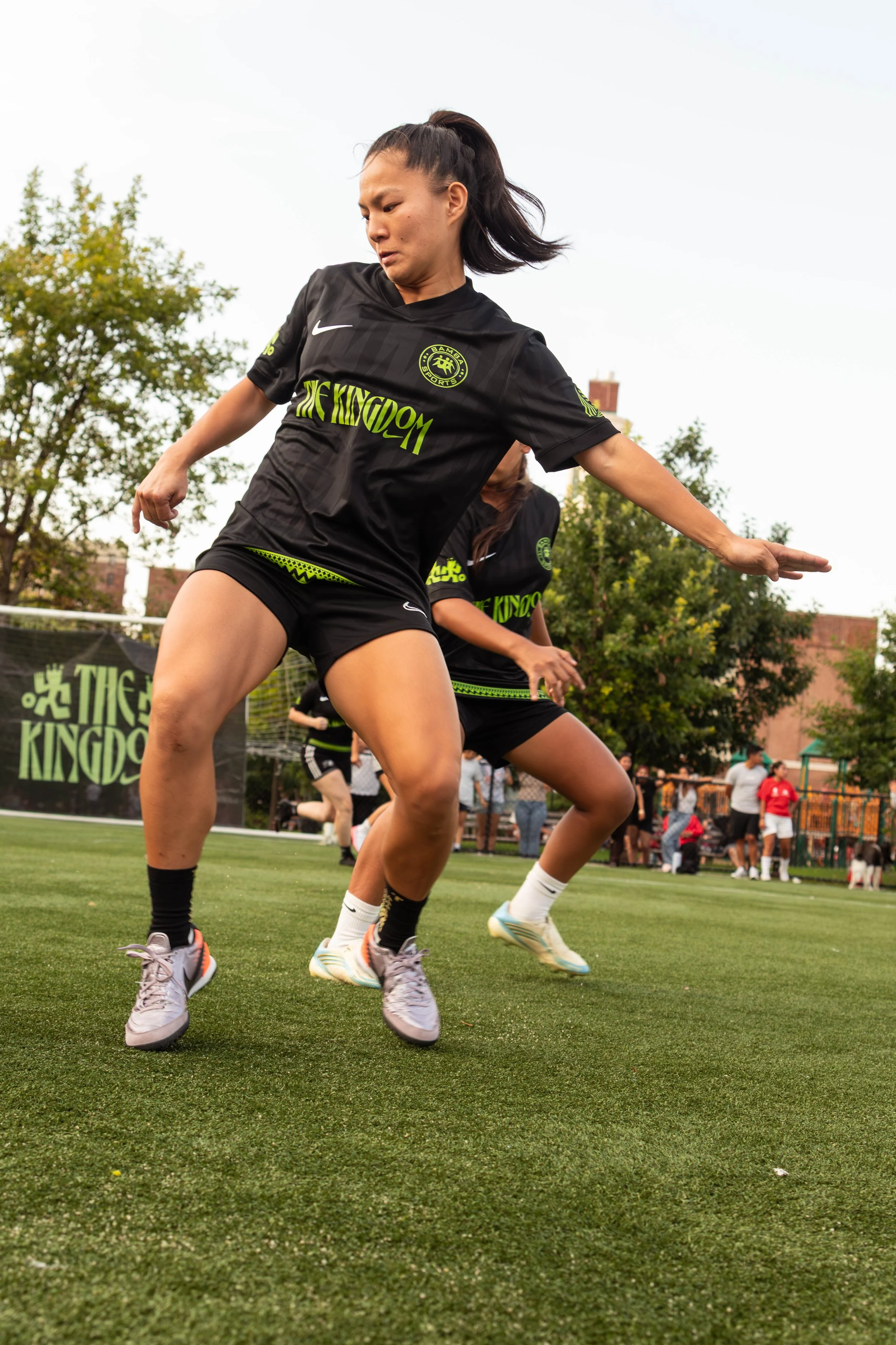 Female soccer players in black uniforms performing a dance or warm-up on a grassy field during a sporting event, with spectators in the background.