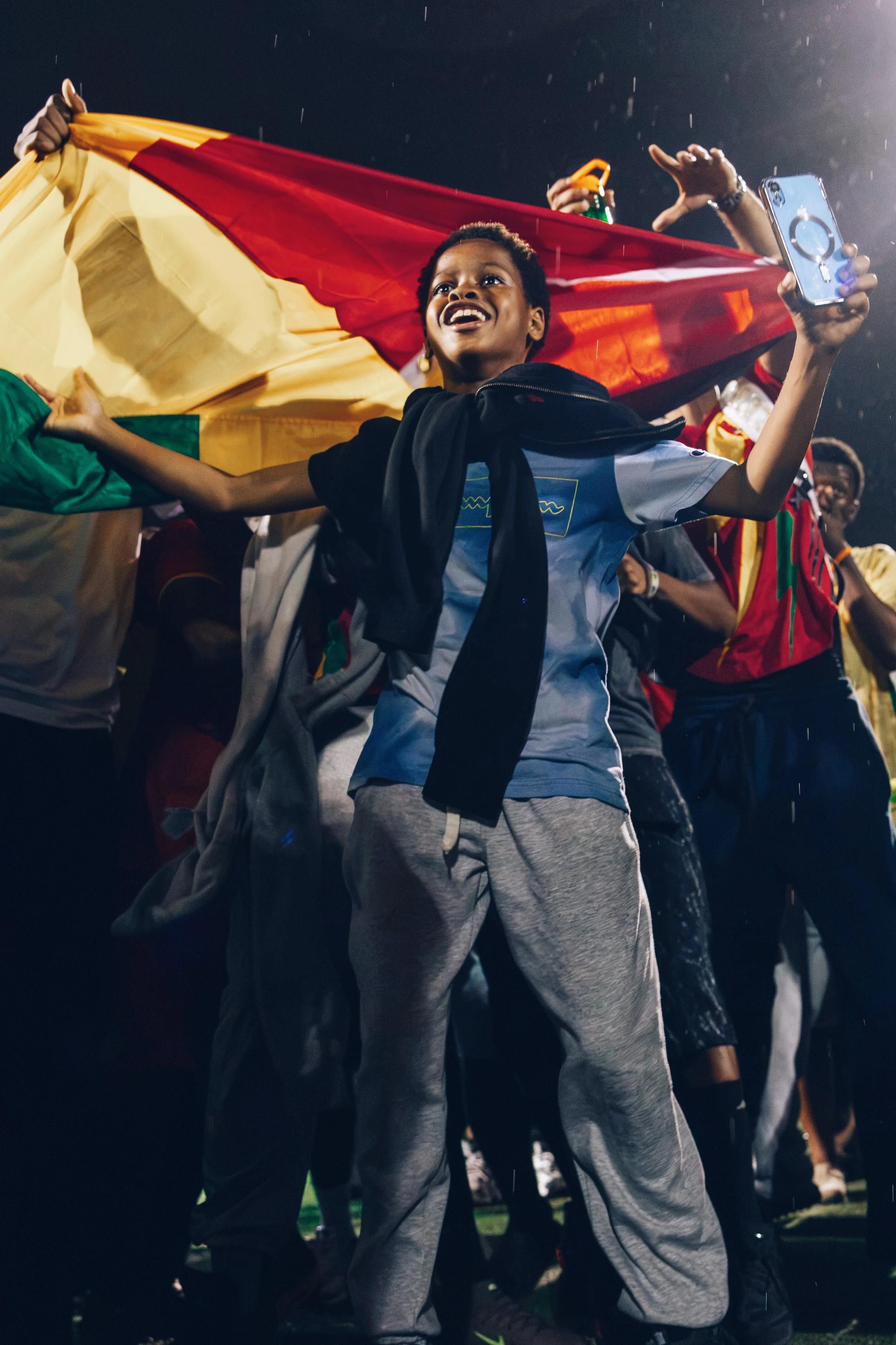 A young person smiling and holding a smartphone with a ring holder, surrounded by a crowd at night, holding a large red and yellow flag.