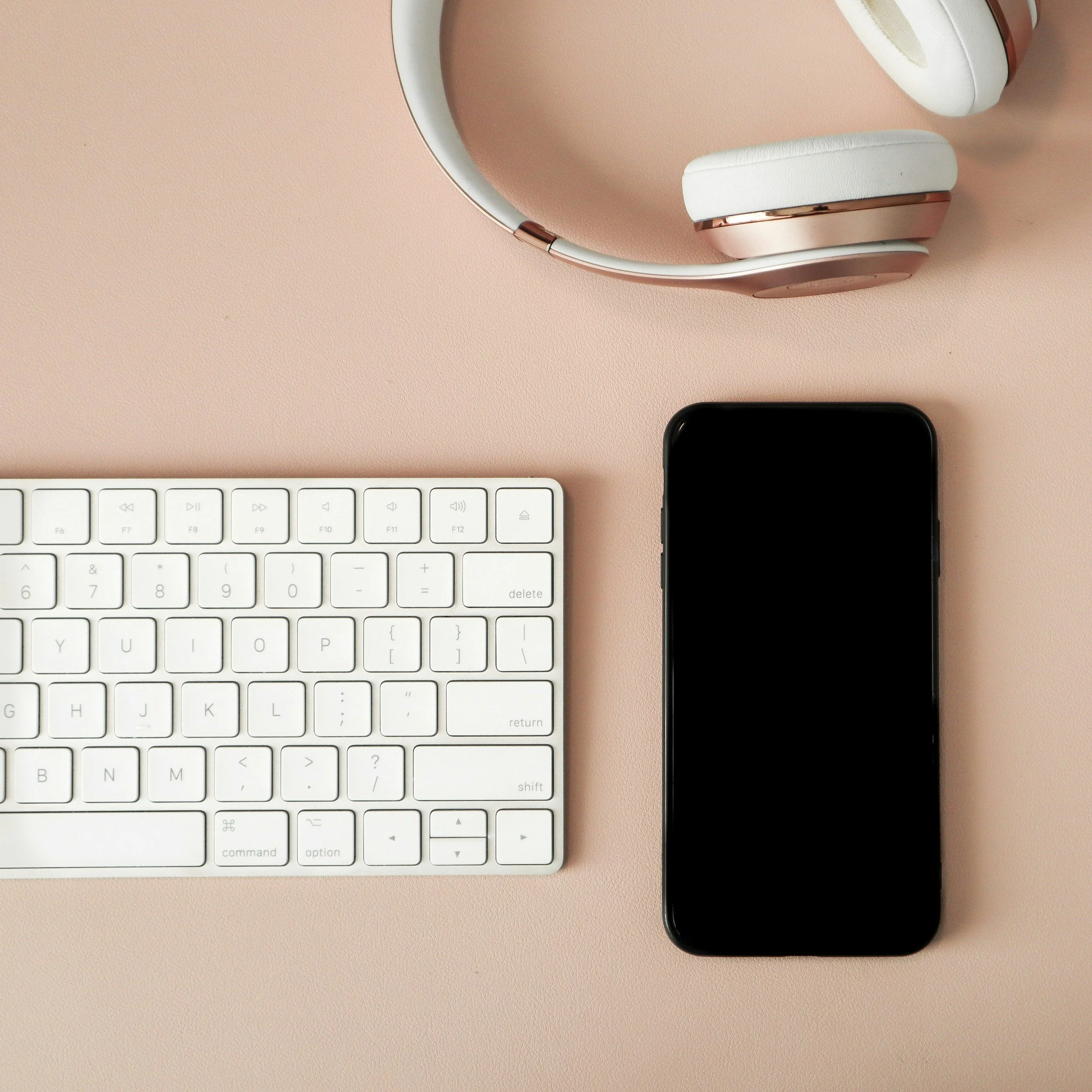 A white computer keyboard, a pair of white and rose gold headphones, and a black smartphone on a pink background.