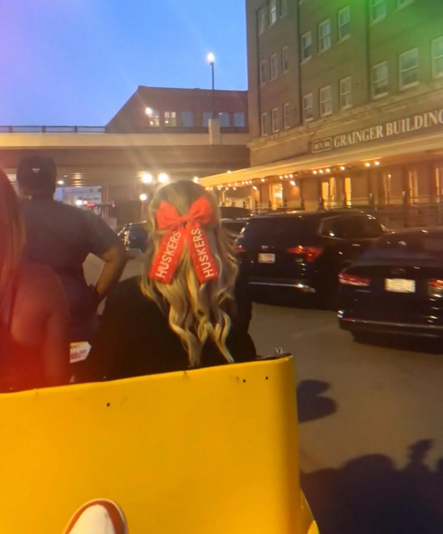 Woman with red 'Huskers' bow in hair, sitting on yellow vehicle at night, near Grainger Building.