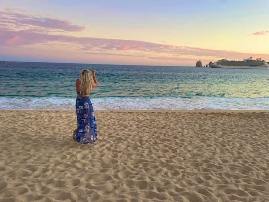 A person standing on a sandy beach with a sunset view over the ocean and a distant cruise ship.