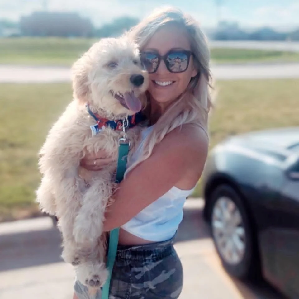 Woman holding a happy, fluffy dog in a parking lot, wearing sunglasses and casual clothes.