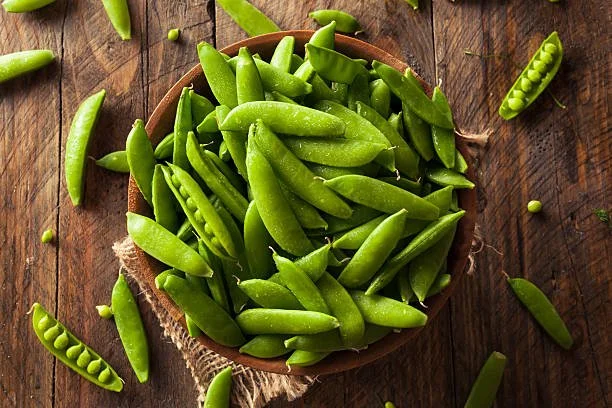 Fresh green snap peas in a wooden bowl on a rustic wooden surface.