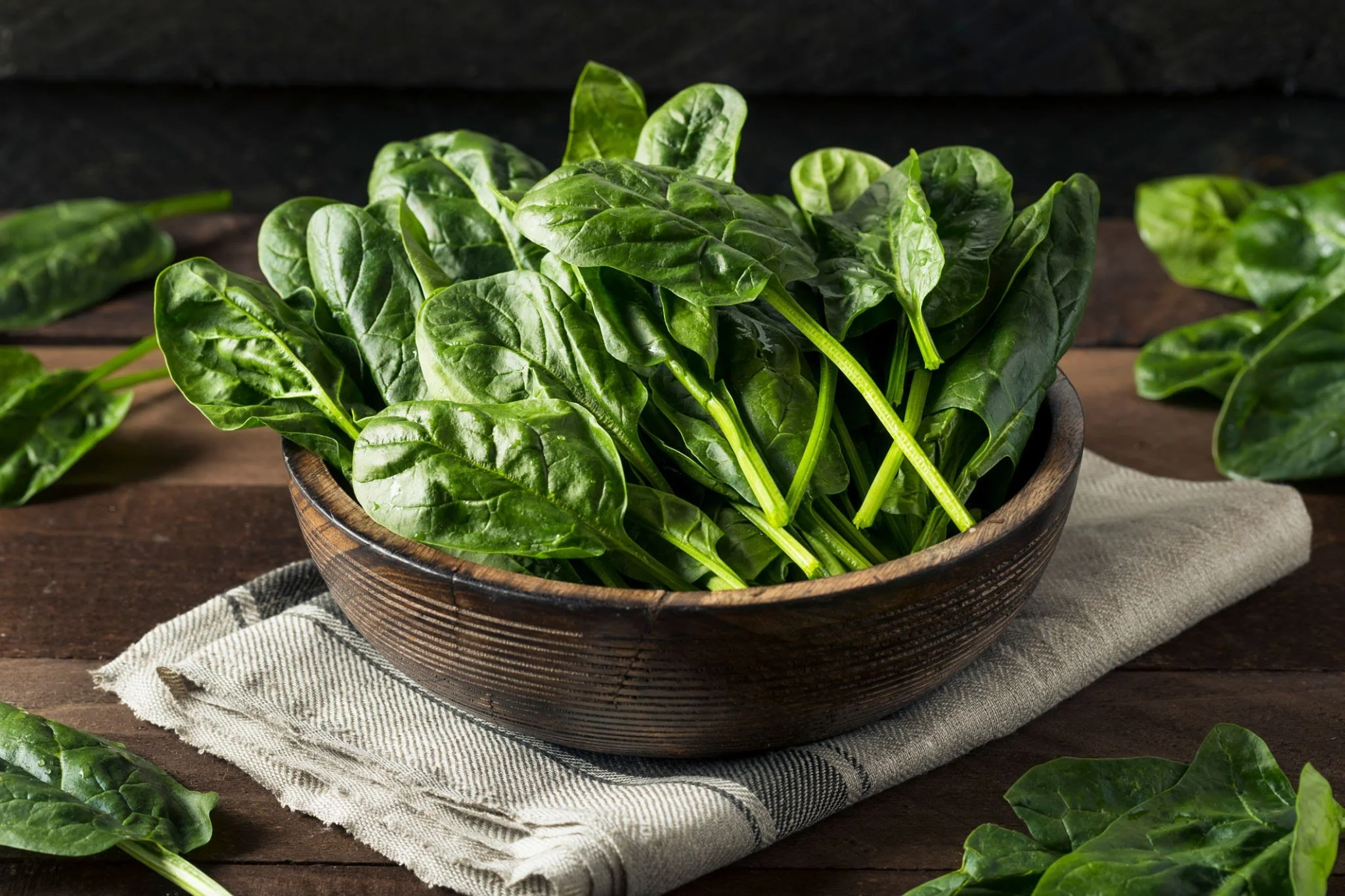Fresh spinach leaves in a wooden bowl on a dark wooden table with a beige cloth underneath.
