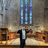 A young person standing inside a church with high stone walls and stained glass windows, holding a book with arms raised.
