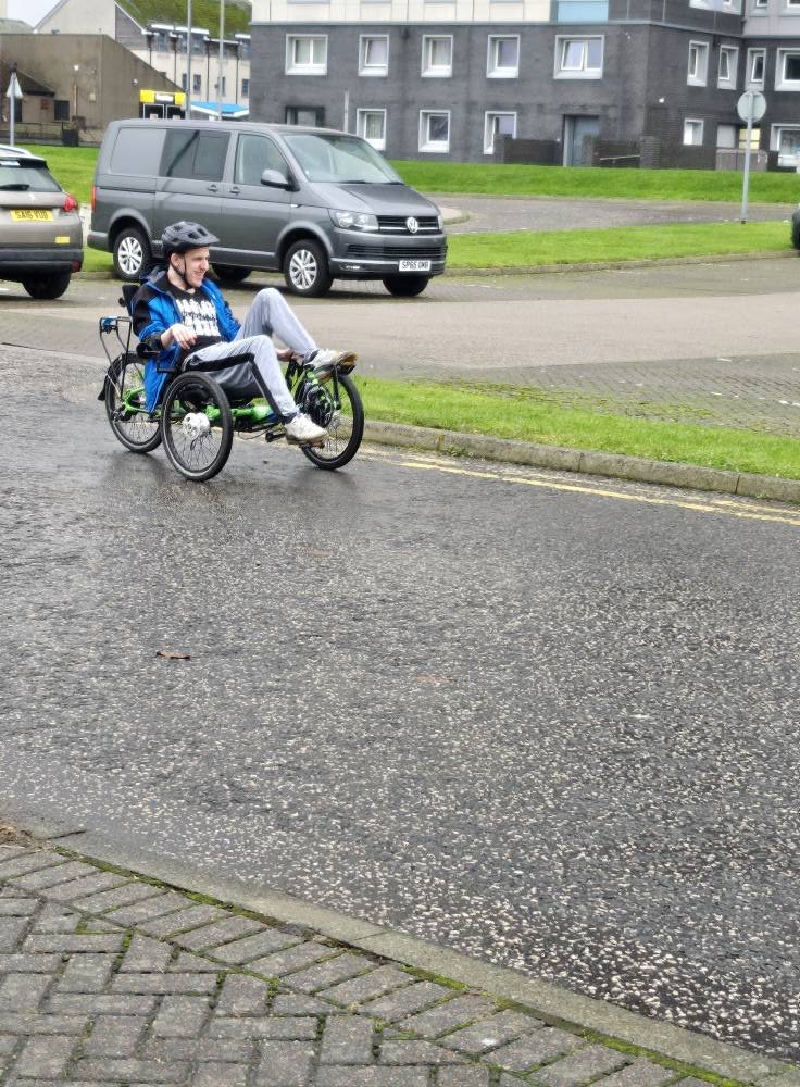 A person riding a recumbent tricycle on a wet street in an urban area, with parked cars and residential buildings in the background.