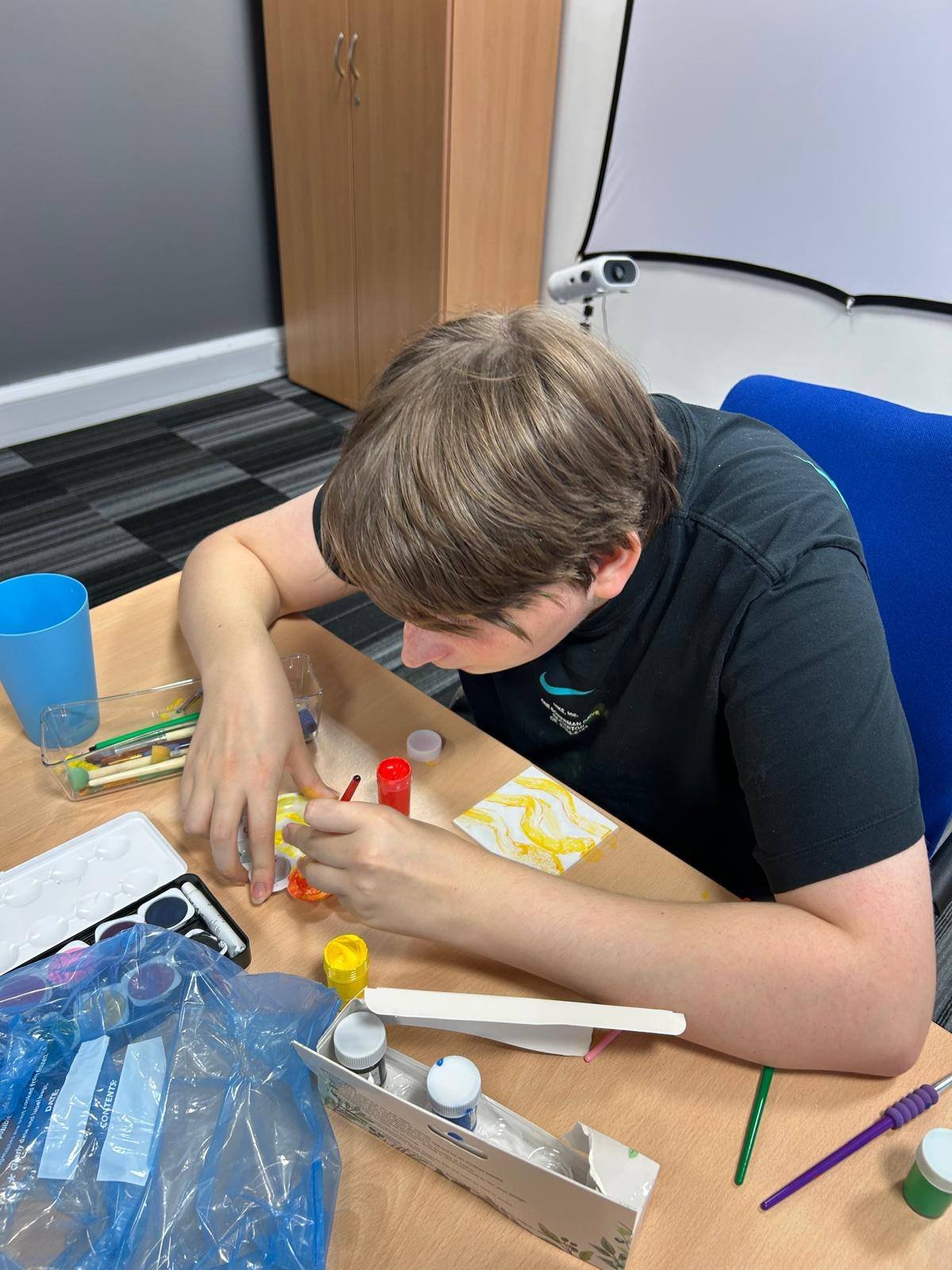 A young boy sitting at a table, engaged in a painting or craft project with various paints, brushes, and supplies around him.