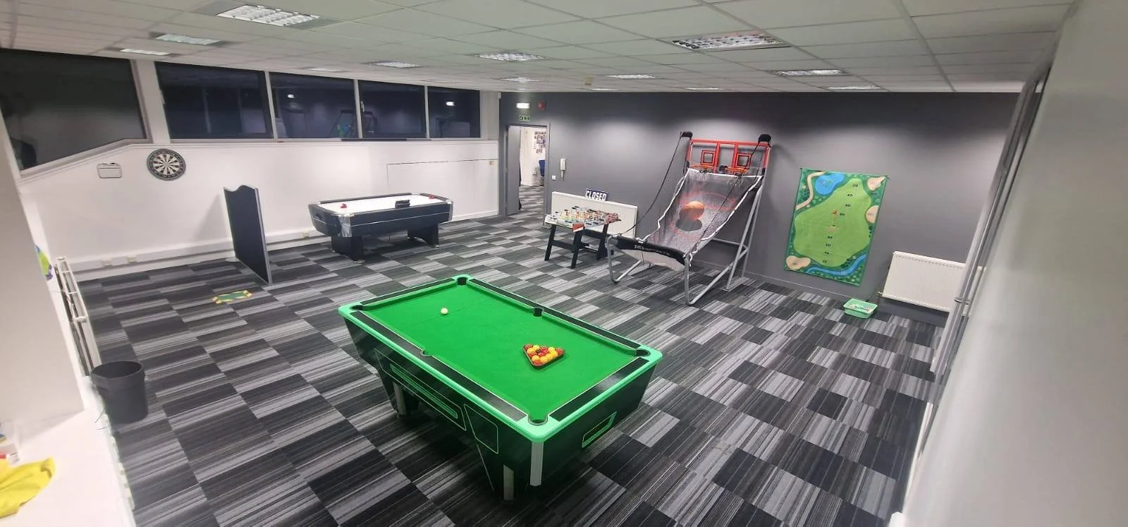 An indoor game room with a pool table, air hockey, basketball arcade game, dartboard, and a miniature golf course setup. The room has gray walls, a tiled gray carpet, and ceiling lights.