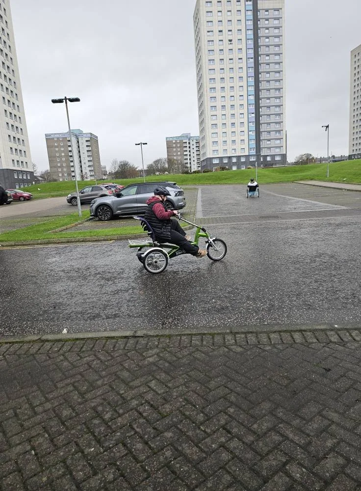 A person riding a recumbent tricycle on a wet parking lot in an urban area with tall apartment buildings in the background.