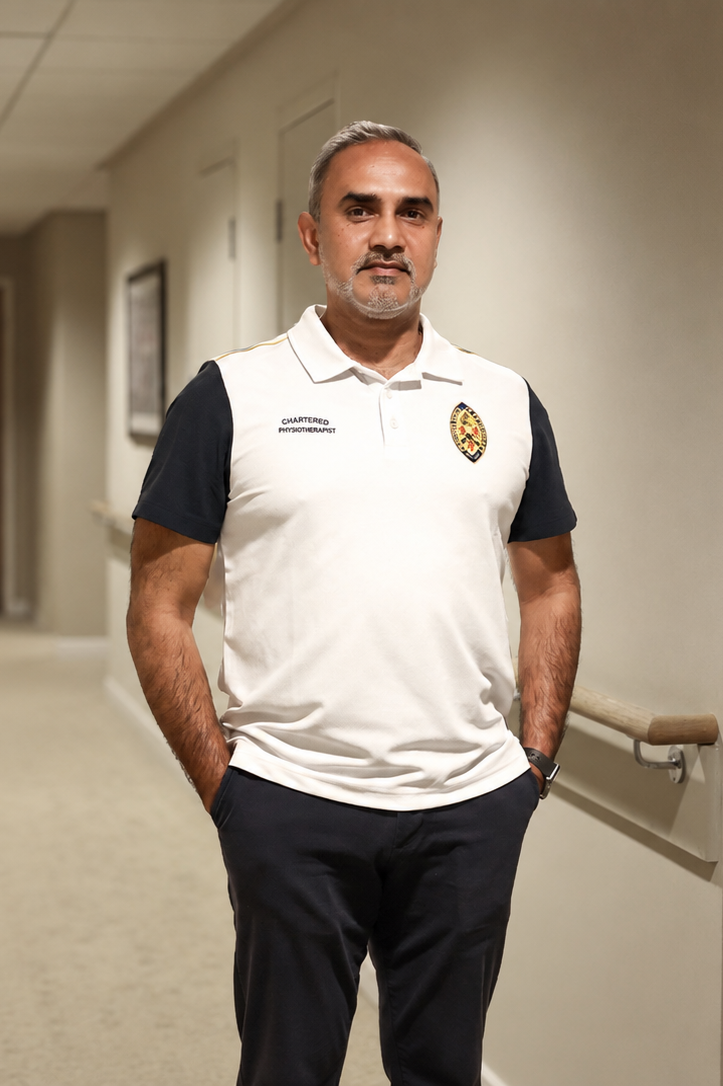A man standing in a hallway wearing a white polo shirt with black sleeves, dark pants, and a wristwatch. The shirt has a logo and the text 'Chartered Physiotherapist' on it.