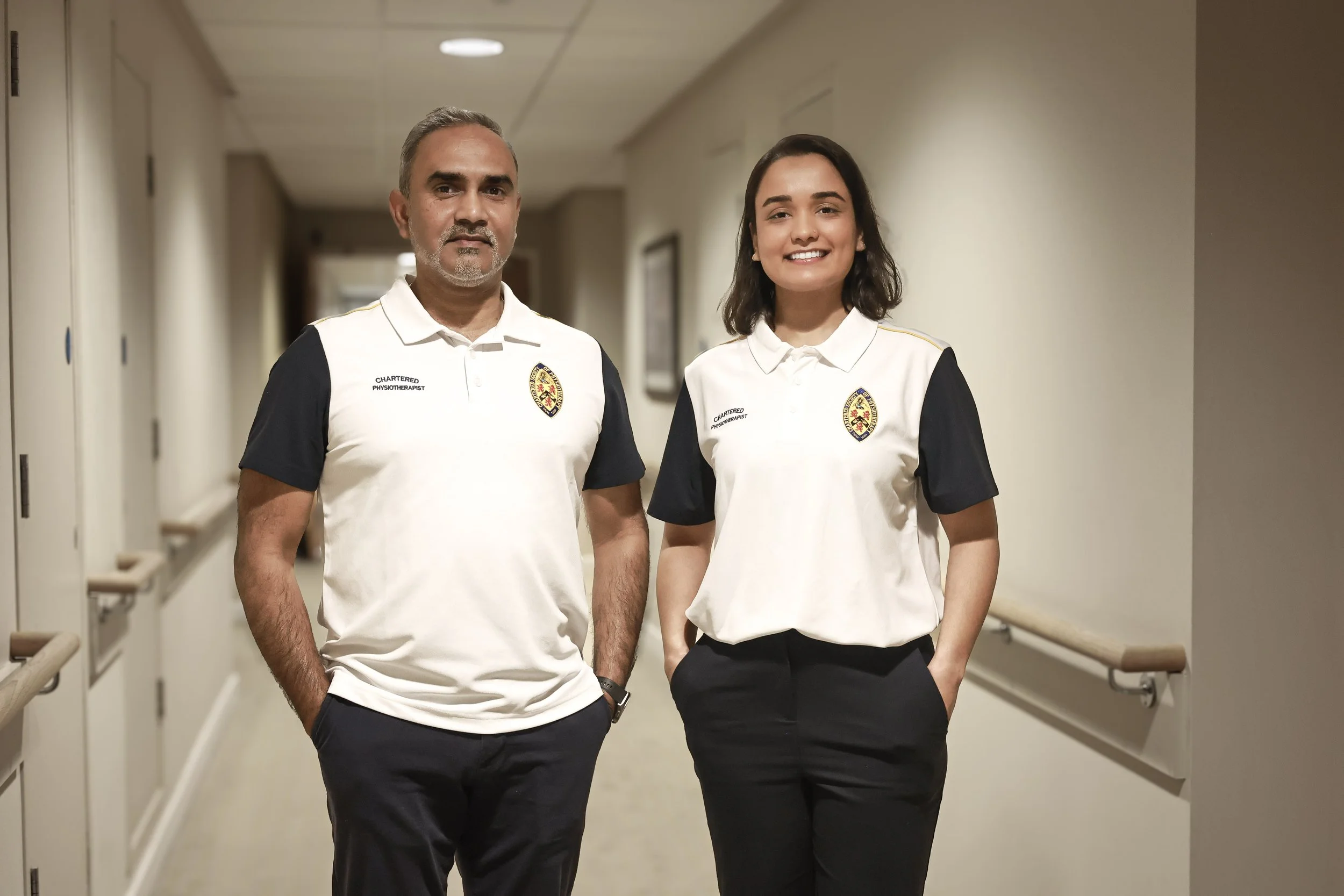 A man and a woman standing in a hospital corridor, both wearing white shirts with a badge and text indicating they are chartered physiotherapists, smiling at the camera.