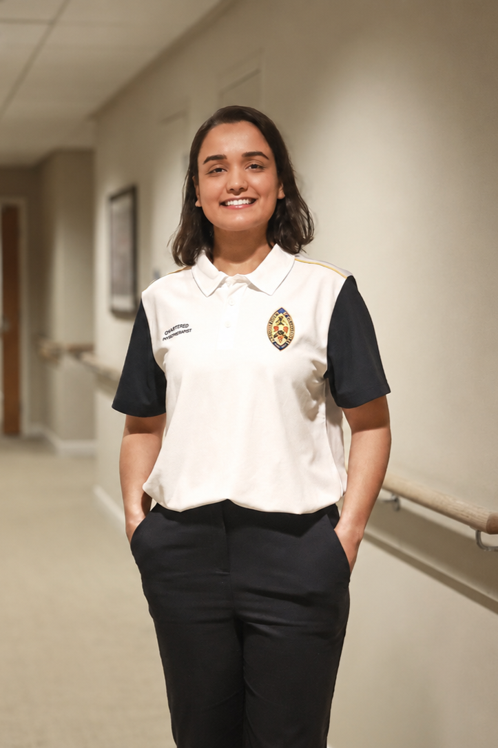 Smiling young woman with shoulder-length dark hair, wearing a white and black uniform with an embroidered badge on the chest, standing in a hallway with hands in pockets.