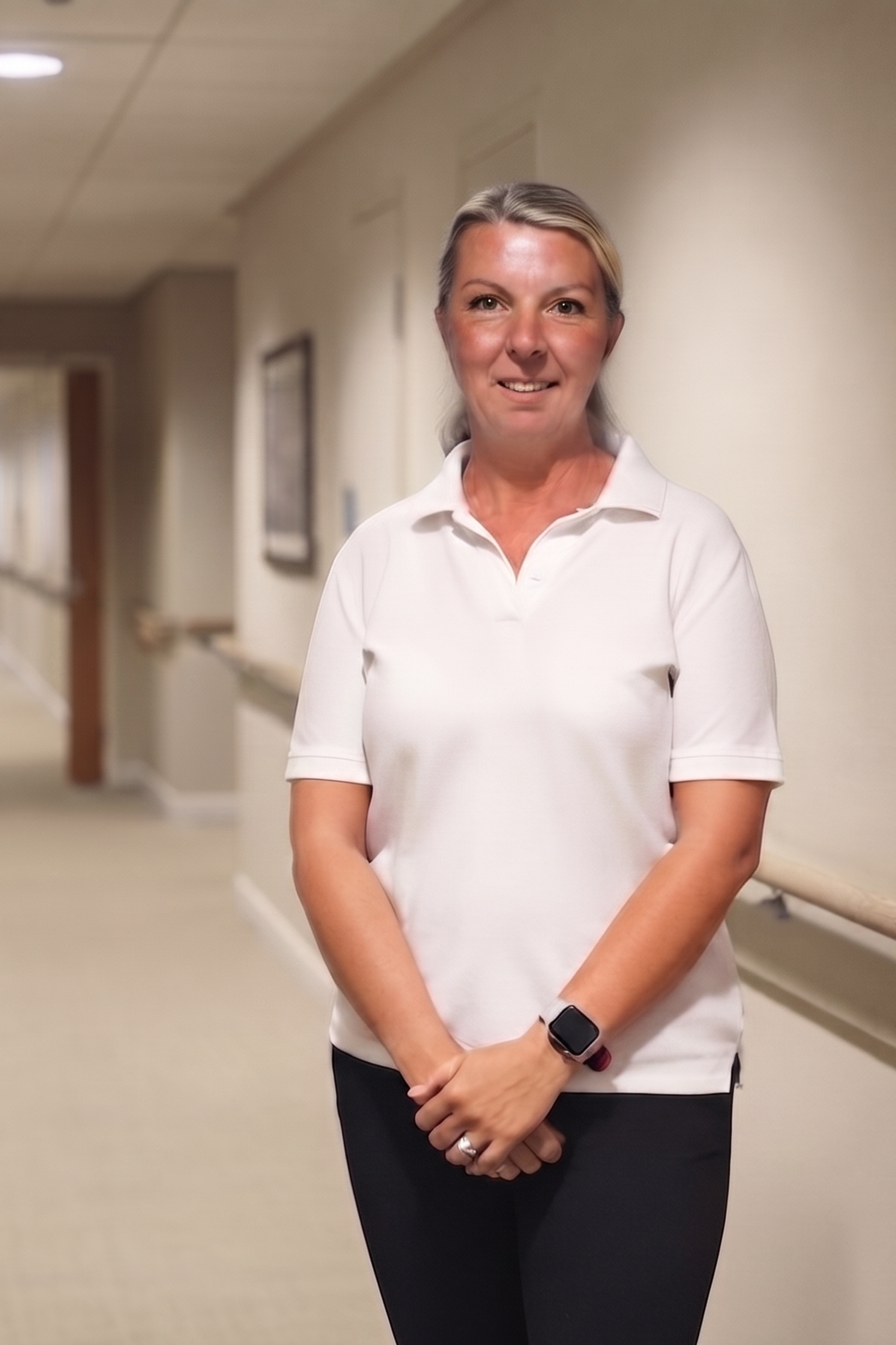 A woman with blonde hair wearing a white polo shirt and black pants standing in a hospital corridor.