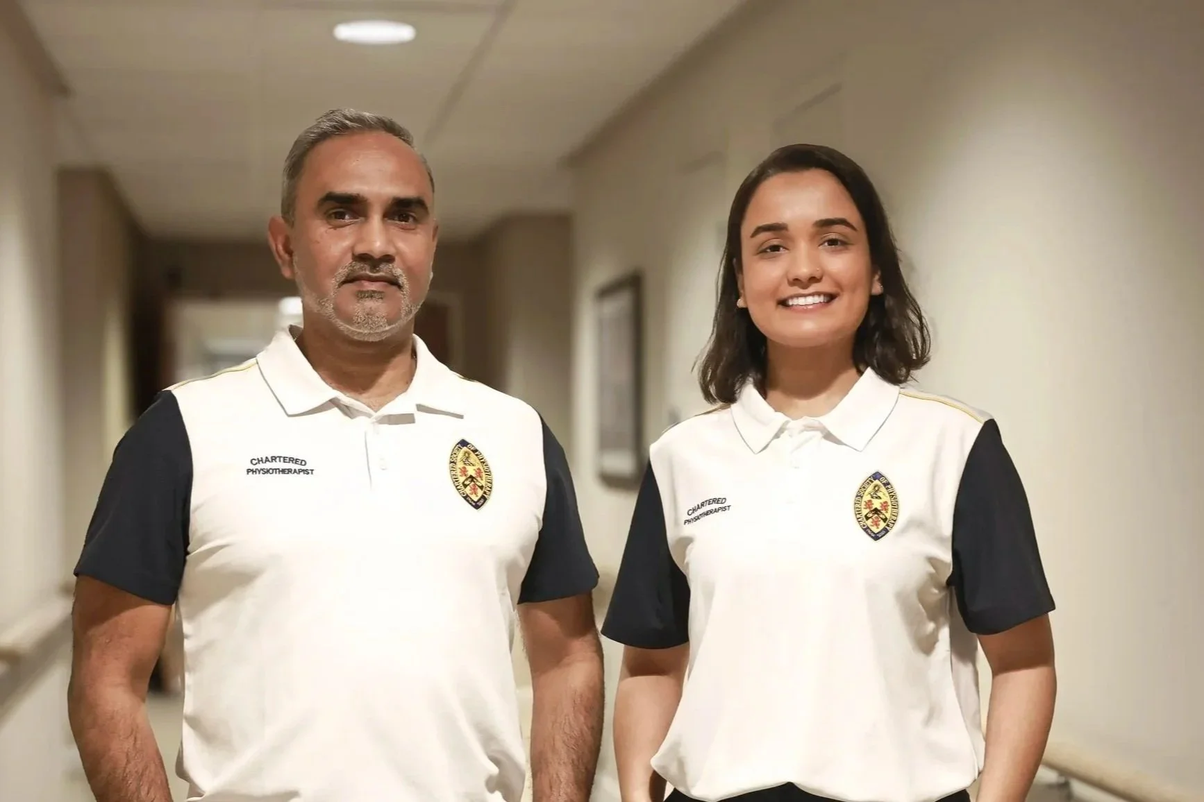 Two individuals, a man and a woman, standing in a corridor, both wearing white and black sports shirts with a badge and the words 'Chartered Physiotherapist' on the shirt, smiling at the camera.