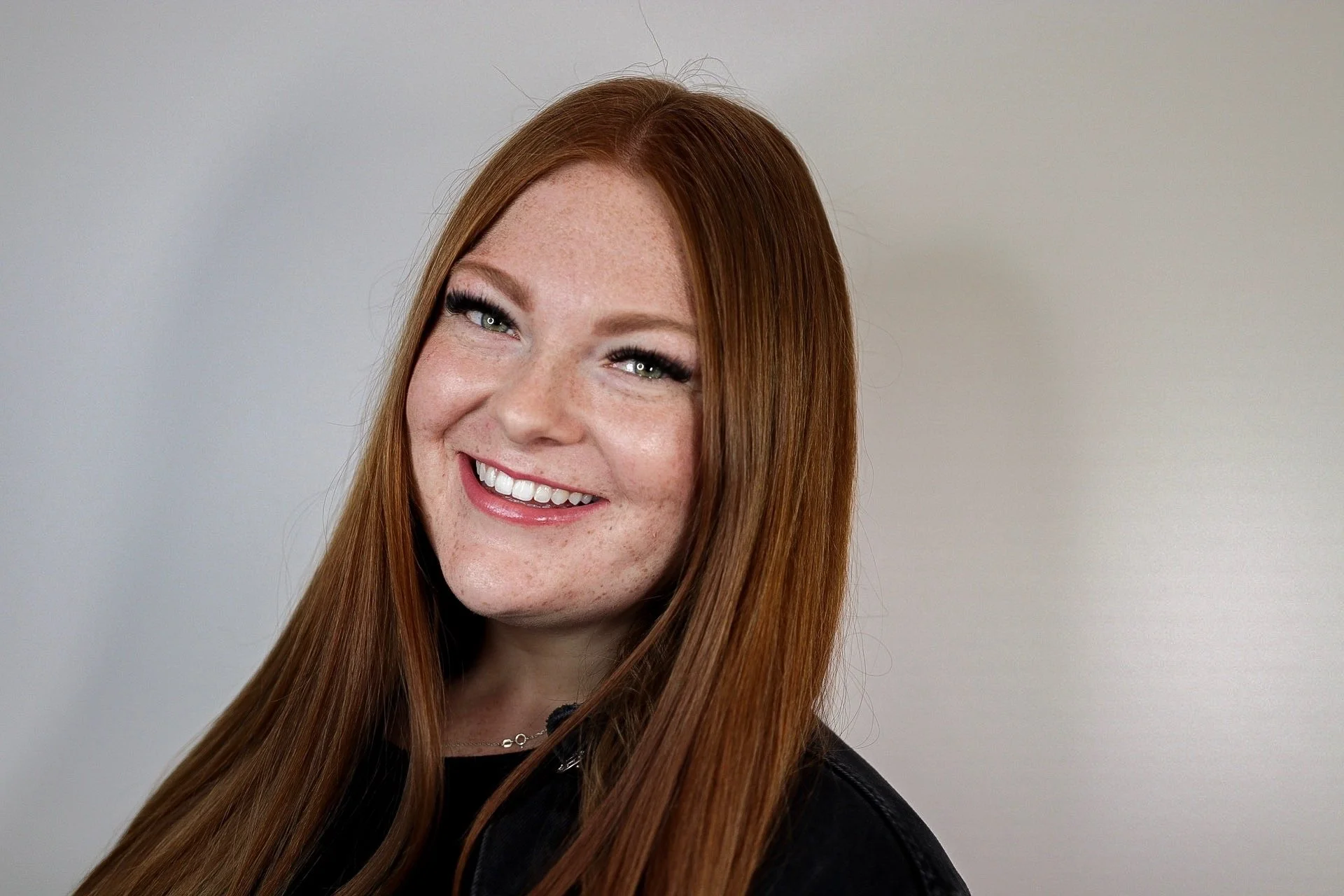 A woman with long red hair smiling, wearing makeup, a black top, and a silver necklace, standing against a plain light-colored background.