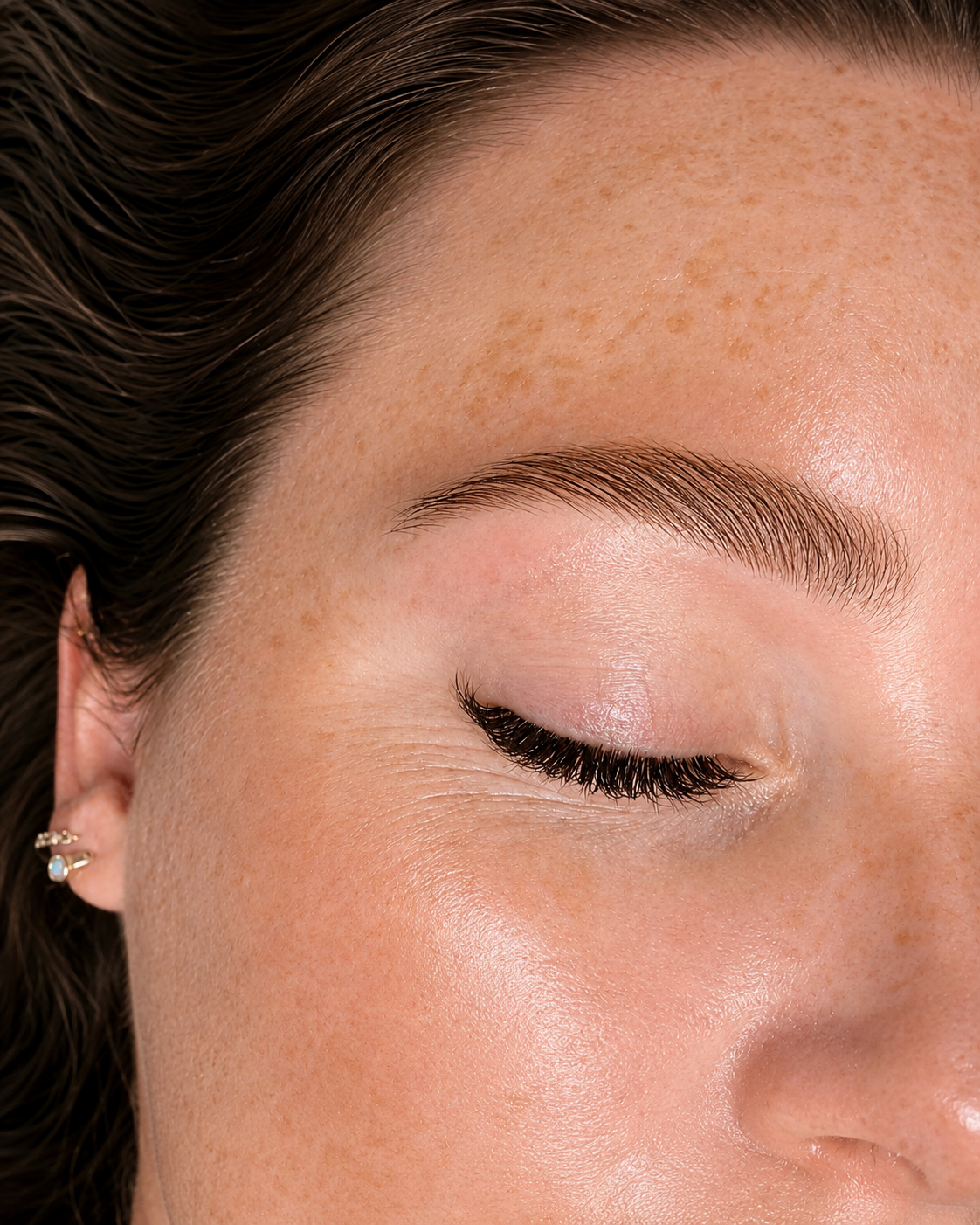 Close-up of a woman's face with closed eye, showing smooth skin, natural freckles, and a well-groomed eyebrow with makeup and mascara.