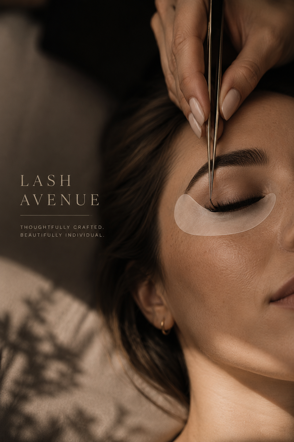 Close-up of a woman receiving eyelash extension treatment with carefully applied false lashes, with eye tape and a professional using tweezers for application. There is a salon environment in the background, and text that reads "Lash Avenue. Thoughtfully crafted. Beautifully individual."