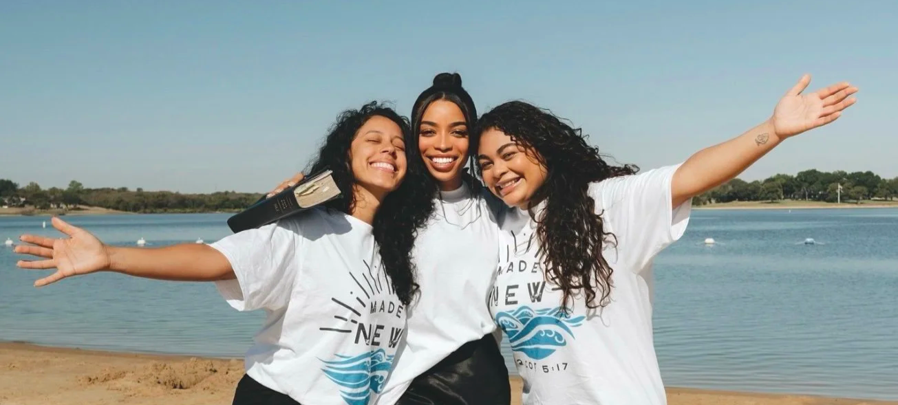 Three smiling women pose together on a sunny beach beside a lake, wearing white graphic T-shirts.