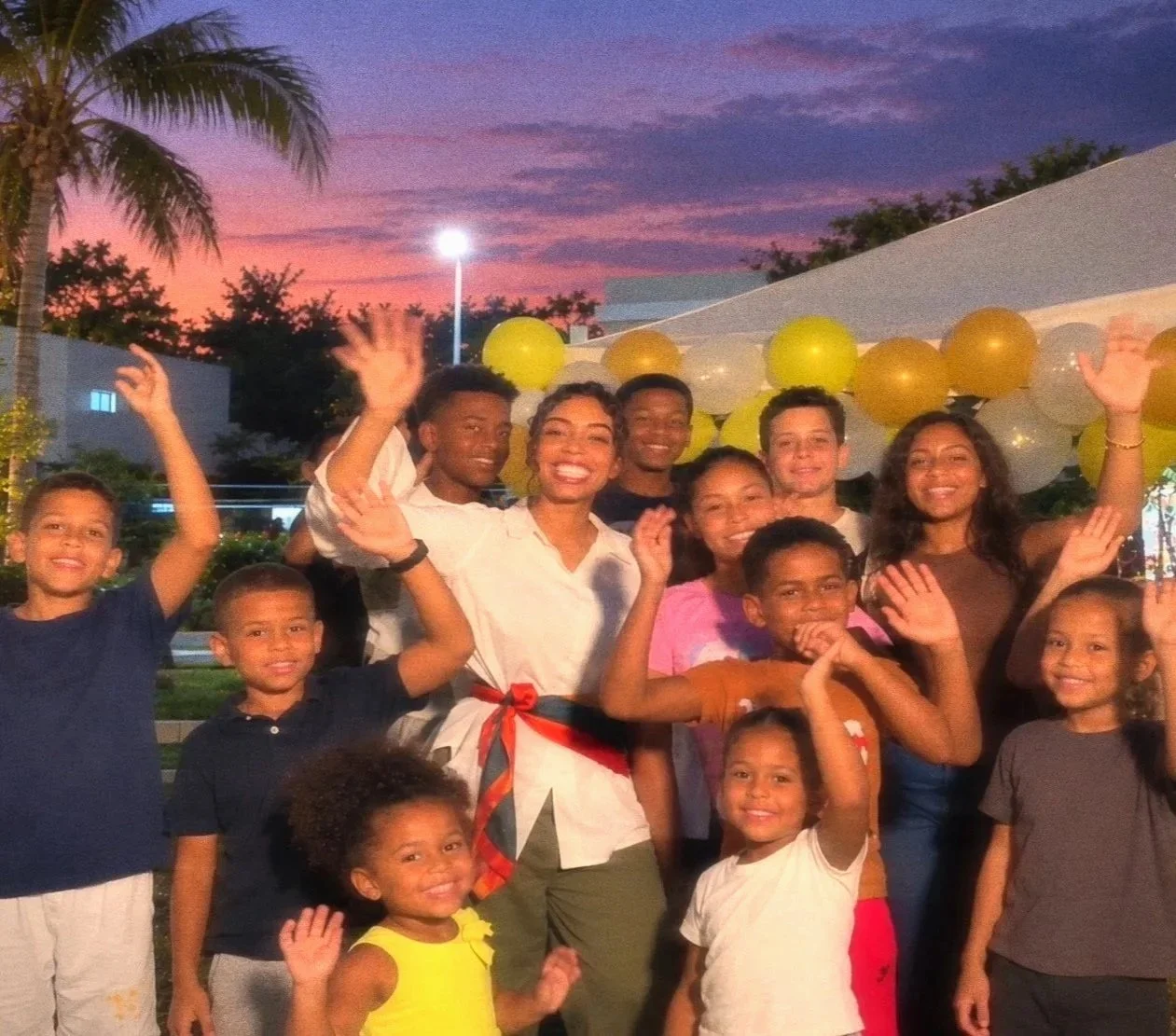 Group of smiling adults and children waving at an outdoor evening party with balloons and palm trees.