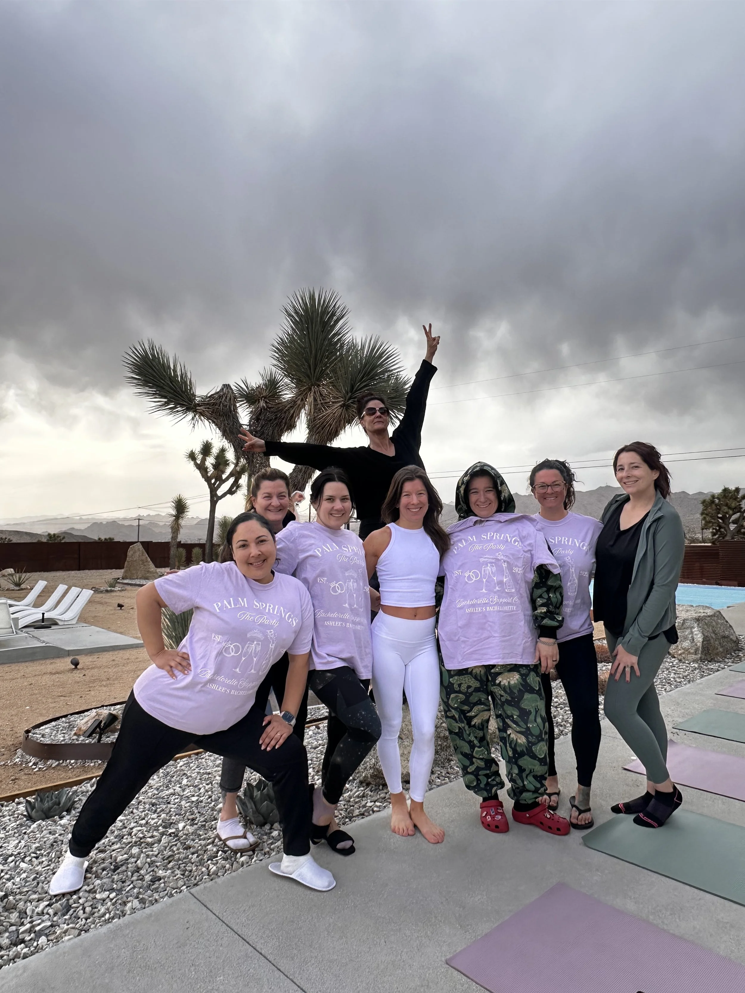 Group of seven women posing outdoors, some wearing matching Palm Springs t-shirts, others in active wear, with yoga mats and desert landscape in the background.