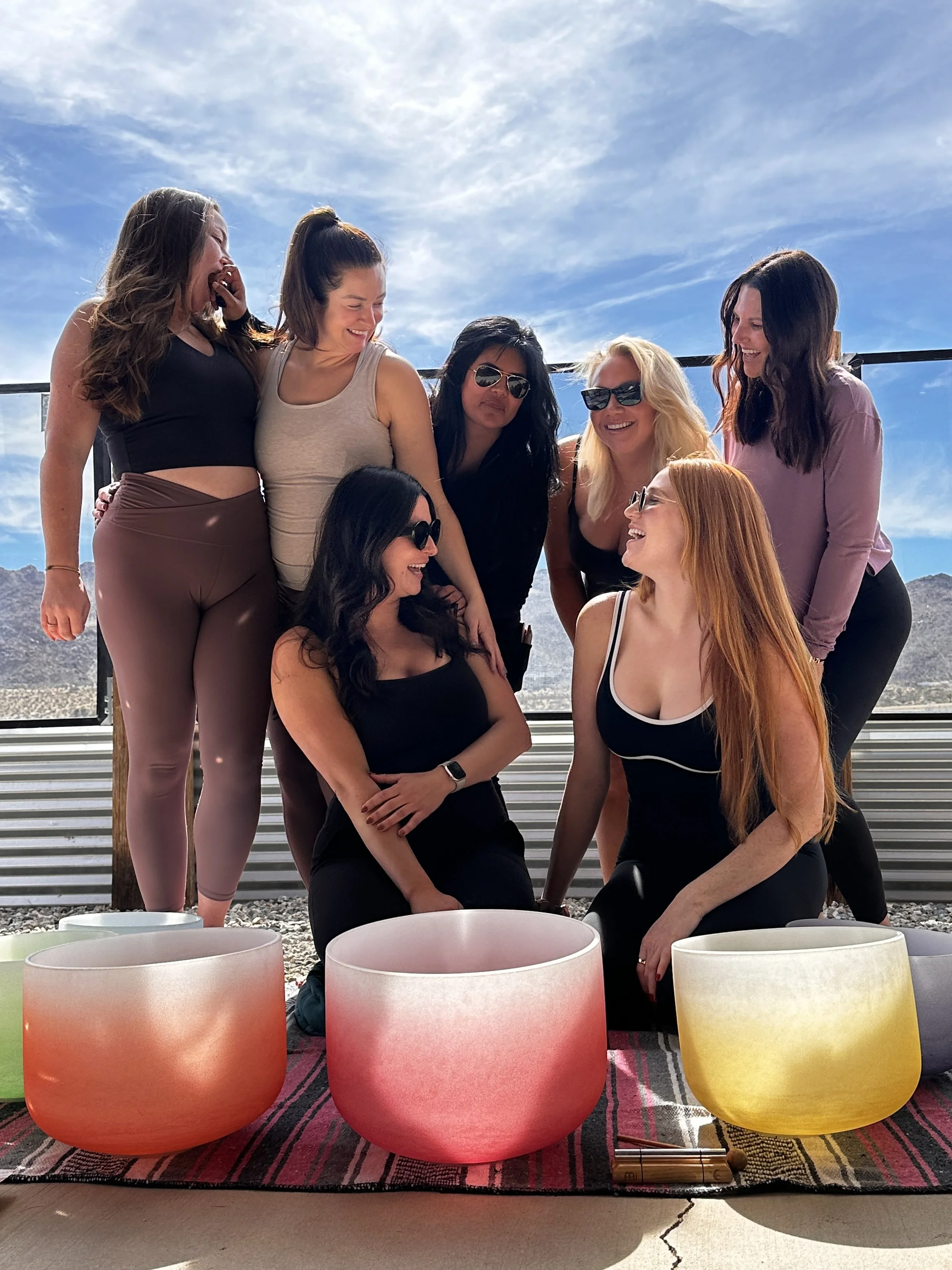 A group of seven women smiling and interacting outdoors, with three colorful singing bowls in the foreground. They are dressed in casual activewear, and there is a blue sky with scattered clouds behind them.
