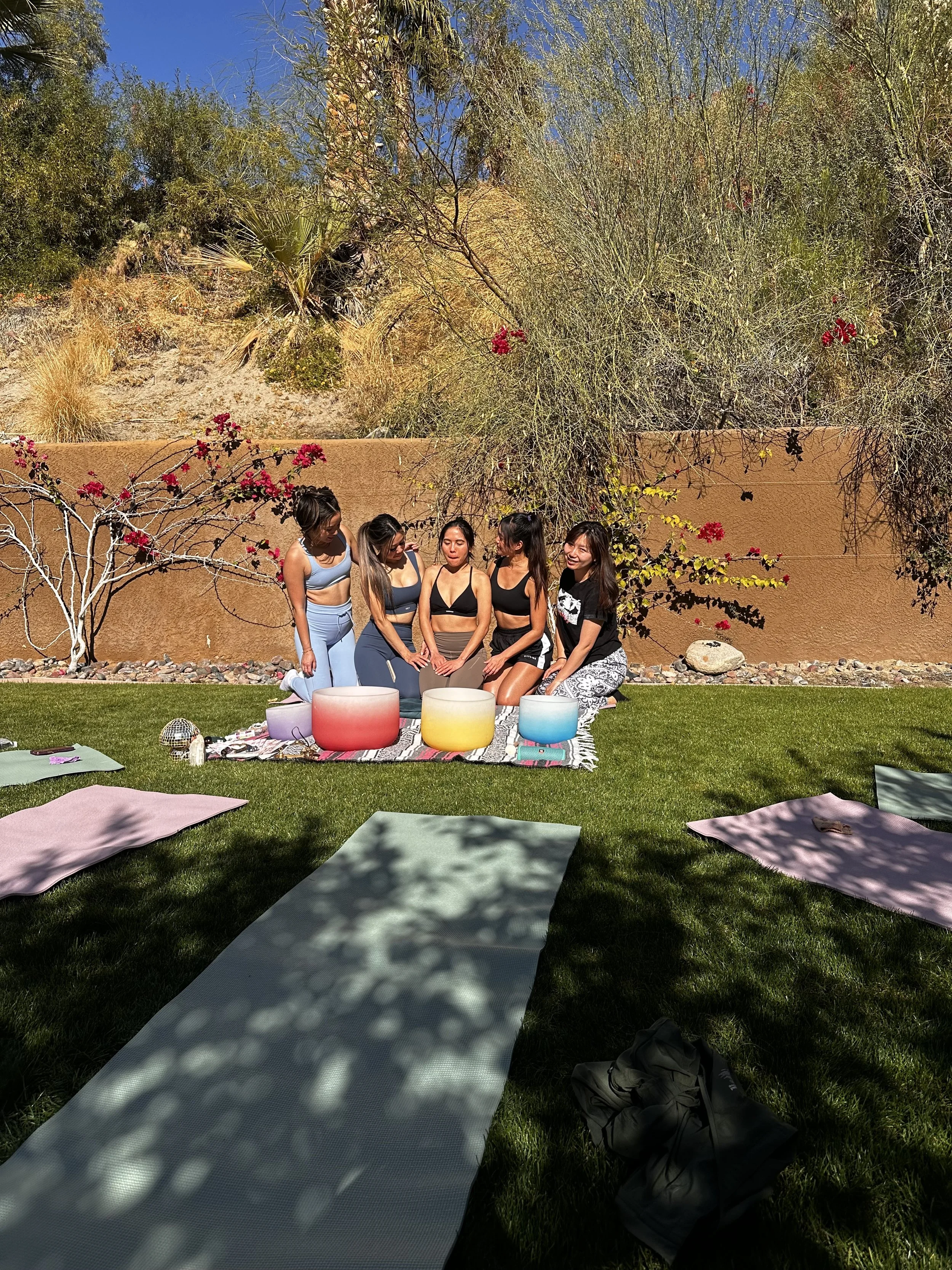 Five women sitting on a grassy area surrounded by yoga mats, wearing workout clothing, with sound bowls in front of them. A desert landscape with shrubs and a wall is in the background.