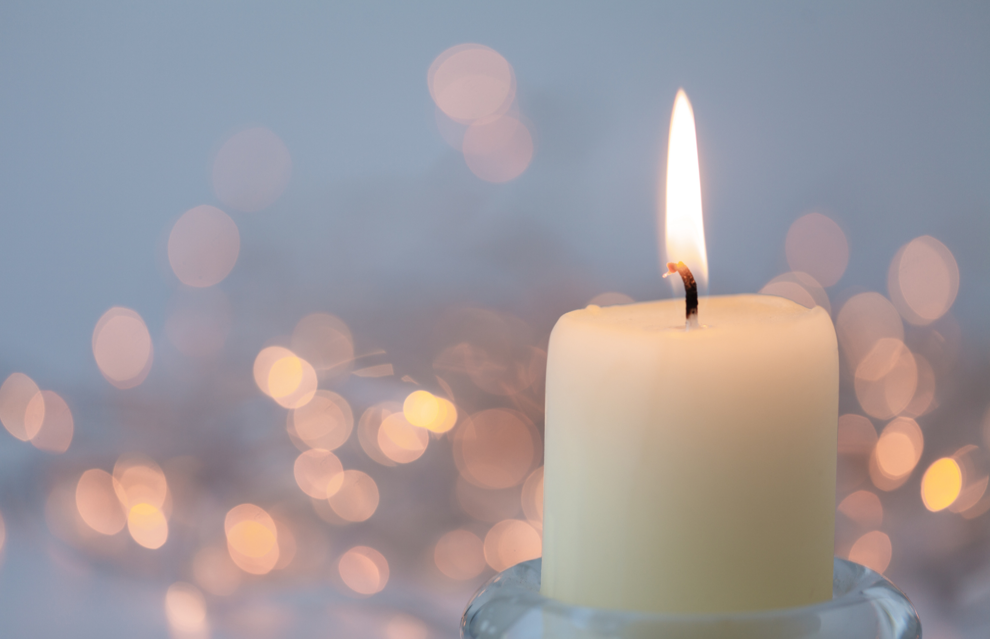 A lit white candle with a glow at the wick, set against a background of blurred lights in warm tones creating a bokeh effect.