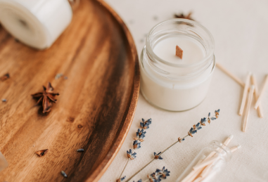 A glass jar candle with a wooden wick, placed on a light-colored surface. Nearby are sprigs of lavender, some clothespins, and a wooden tray.