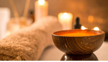 Spa setting with lit candles, a rolled towel, and a wooden bowl on a massage table.