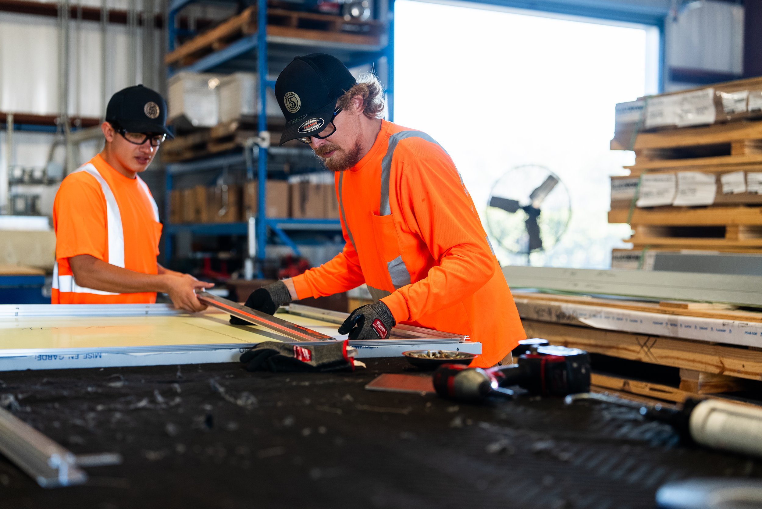 Two workers wearing orange high-visibility shirts and safety hats cutting materials in a warehouse.