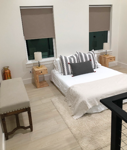 Bedroom with a bed, two wooden nightstands, lamps, a bench, and two windows with gray blinds.