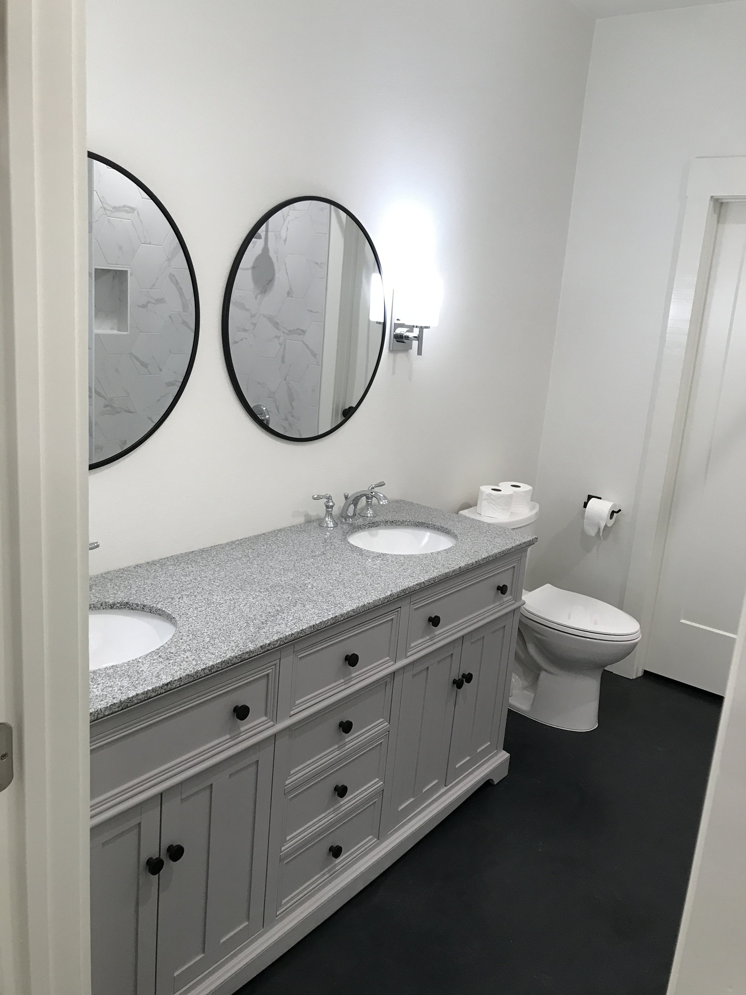 A bathroom with a double sink vanity with gray cabinets, black handles, and a speckled gray countertop. Two oval mirrors are above the sinks, and there is a wall-mounted light fixture between them. Behind the vanity, there's a toilet with a toilet pa