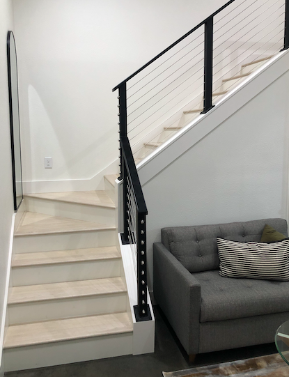 Interior view of a modern staircase with light-colored wooden steps, black metal railing, and a gray sofa with pillows in a living room.