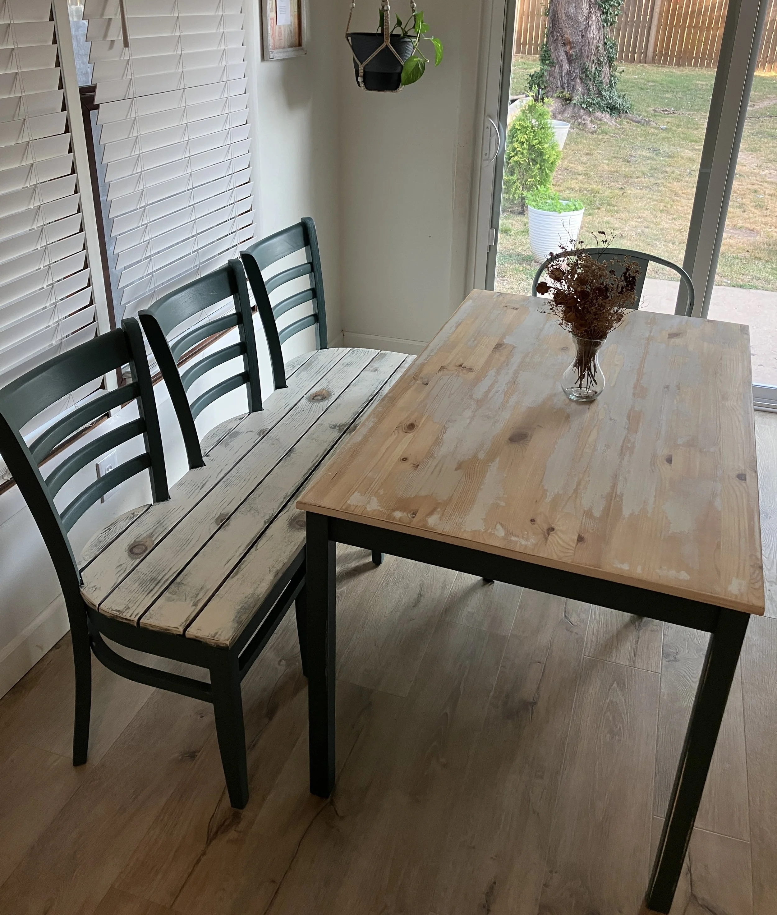 A dining area with a rustic wooden table, four black and gray chairs, a glass vase with dried flowers, white vertical blinds, hanging potted plant, and a sliding glass door that opens to an outdoor yard with grass, trees, and a wooden fence.