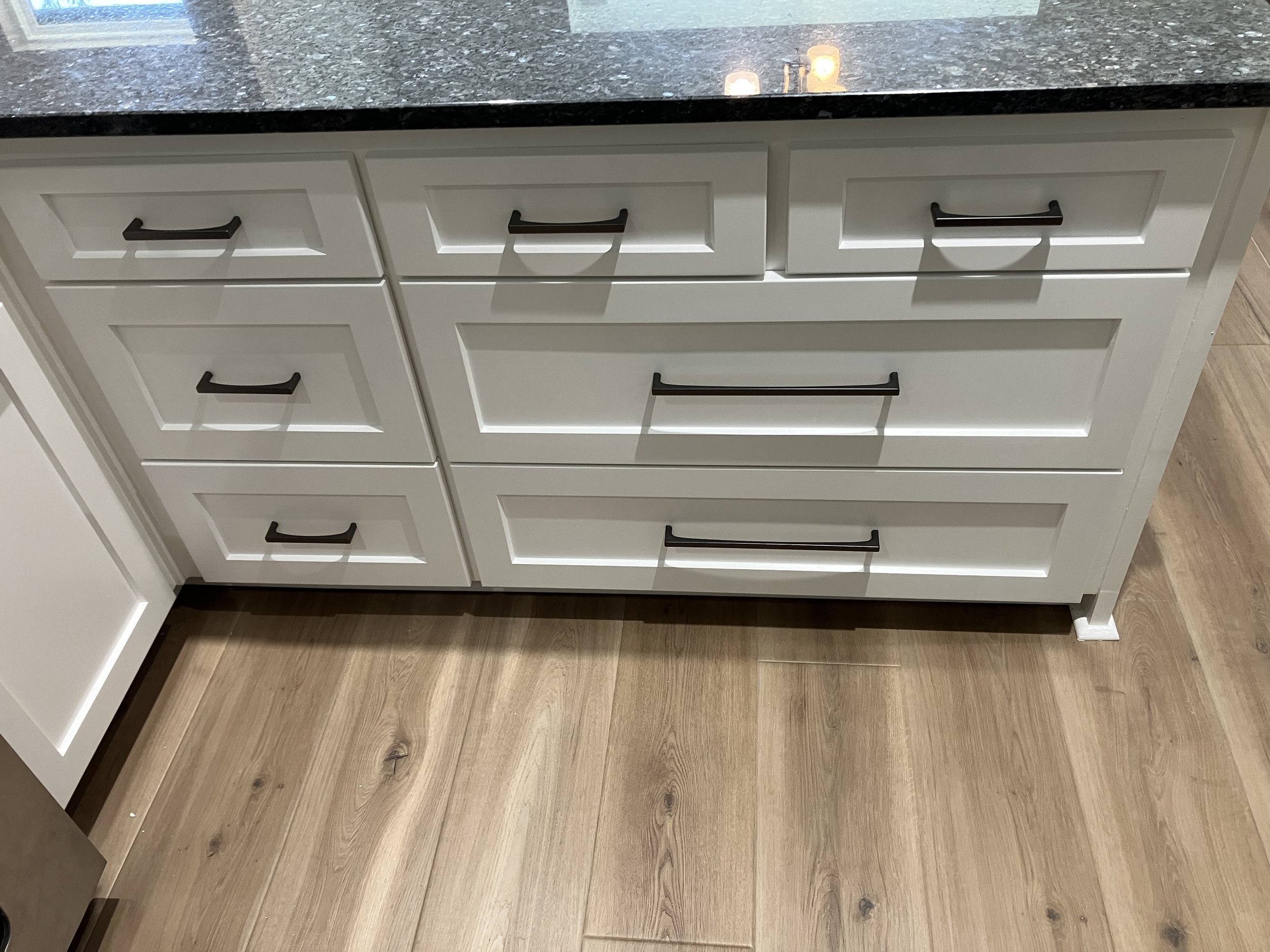 White kitchen cabinets with black handles and a dark granite countertop. The floor is wood with visible knots.