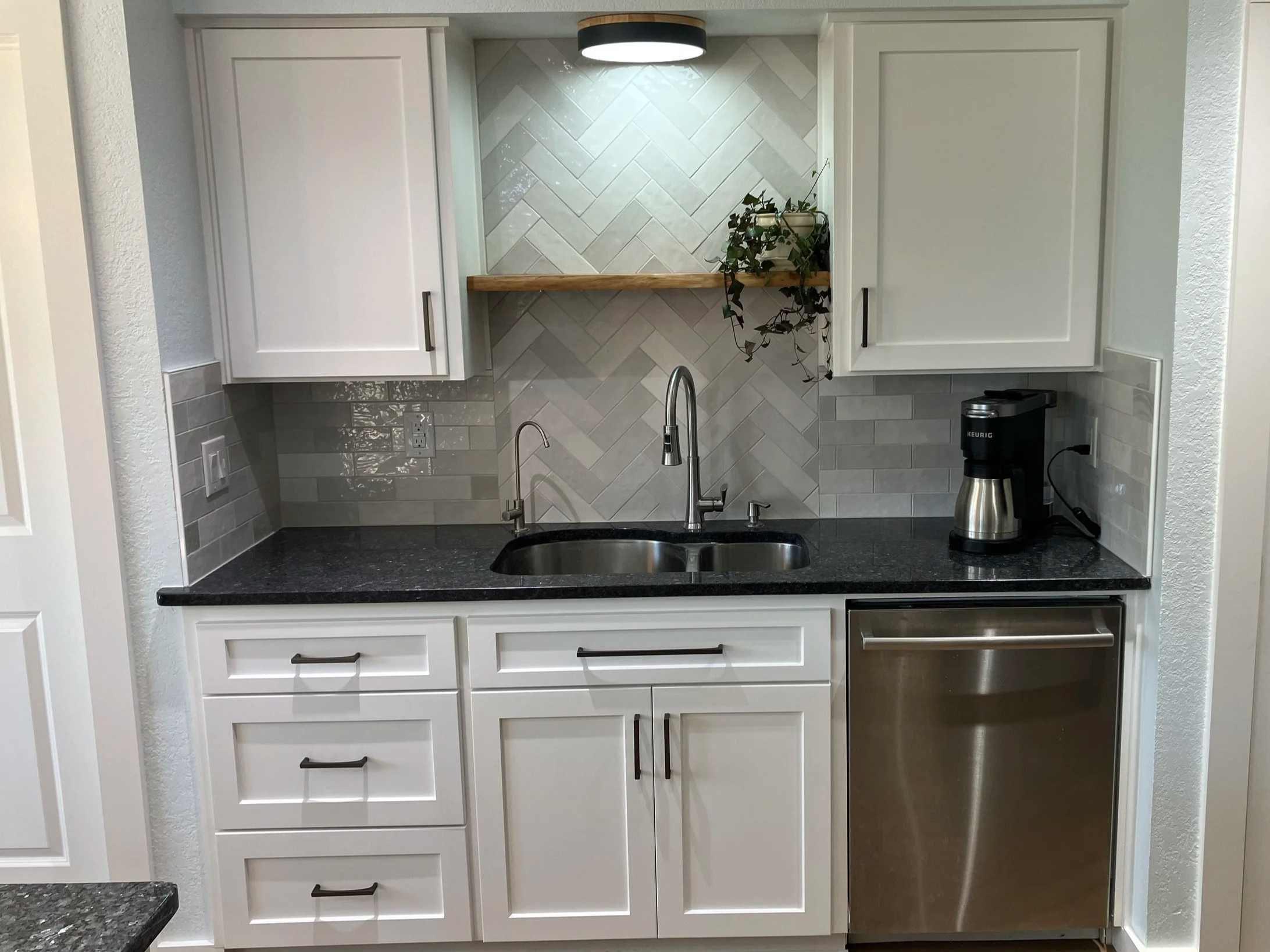 A kitchen counter featuring a dark granite countertop, with white cabinets and drawers below, a stainless steel sink with a modern faucet, a coffee maker, and a silver trash can. Behind the counter, there is a backsplash with diagonal gray tiles and 
